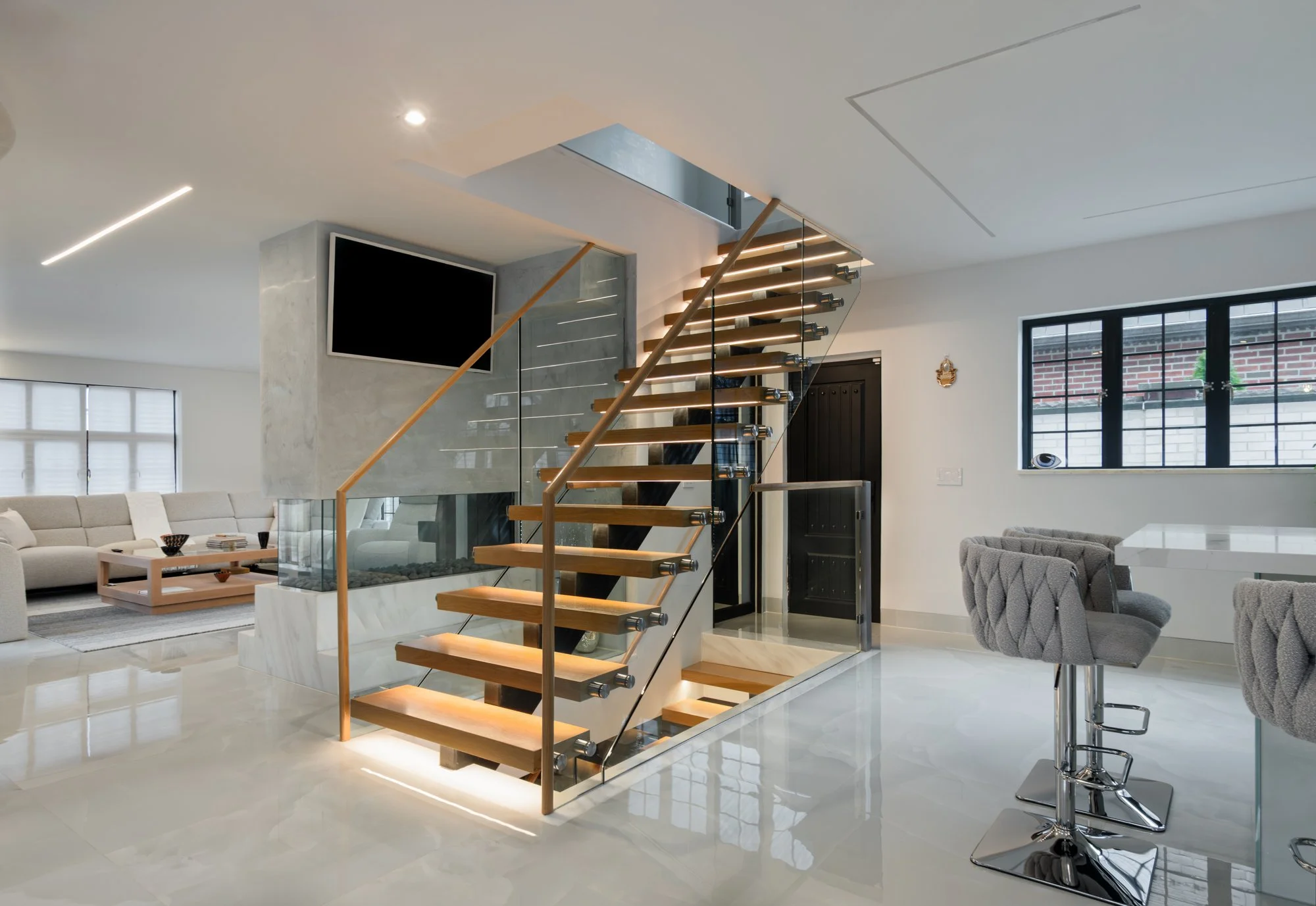Modern minimalistic interior with wooden flooring, staircase with glass railing, open kitchen with white cabinets, and large windows.