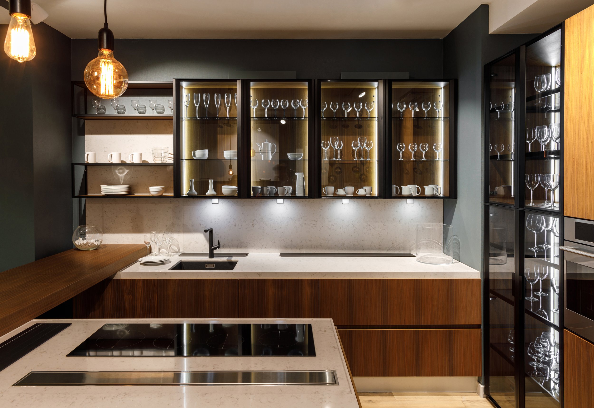 Modern kitchen with wooden cabinets, glass-fronted cupboards, a marble countertop, induction cooktop, and pendant lighting. The cupboards display glassware, cups, and plates.