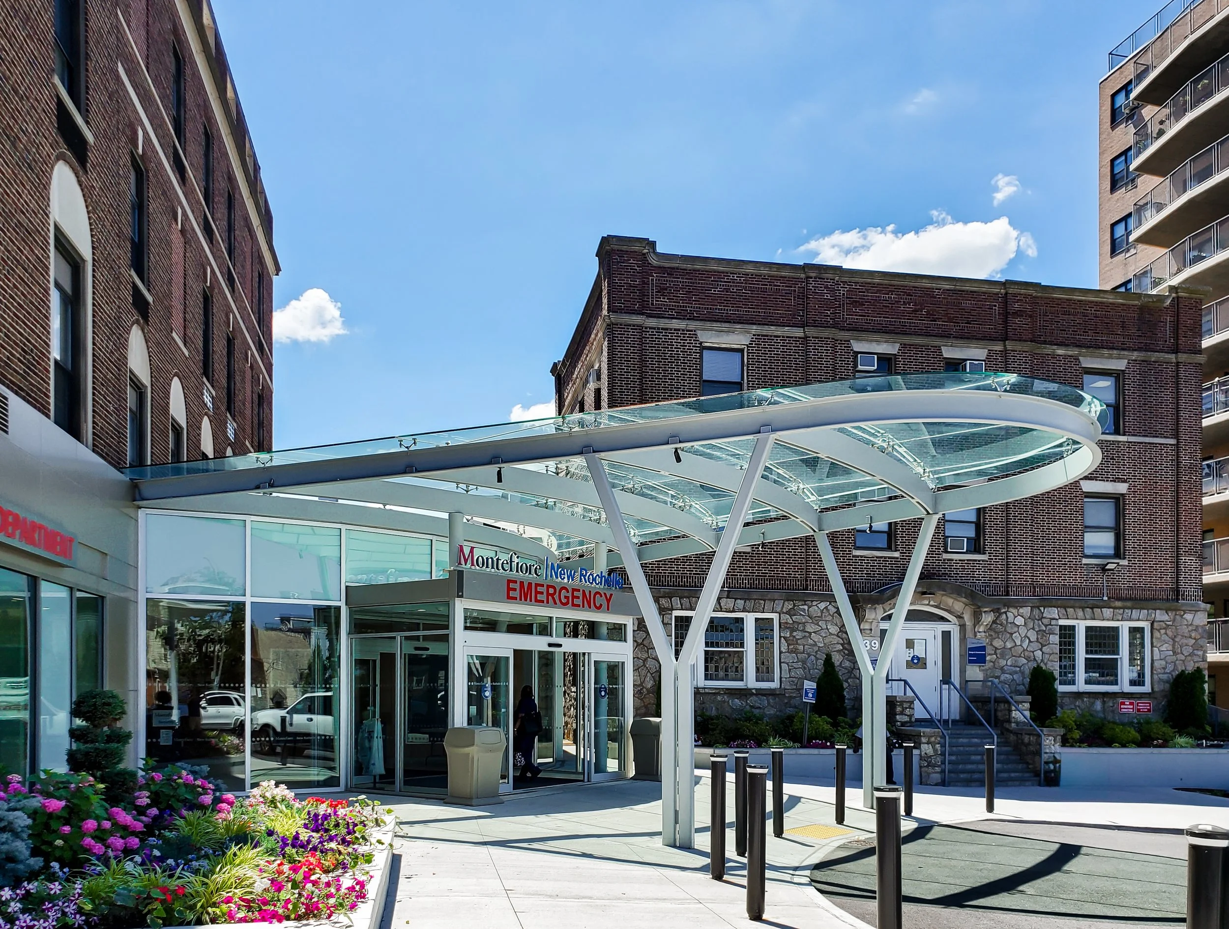 Entrance of a hospital's emergency department with a modern glass canopy and surrounding landscaping.