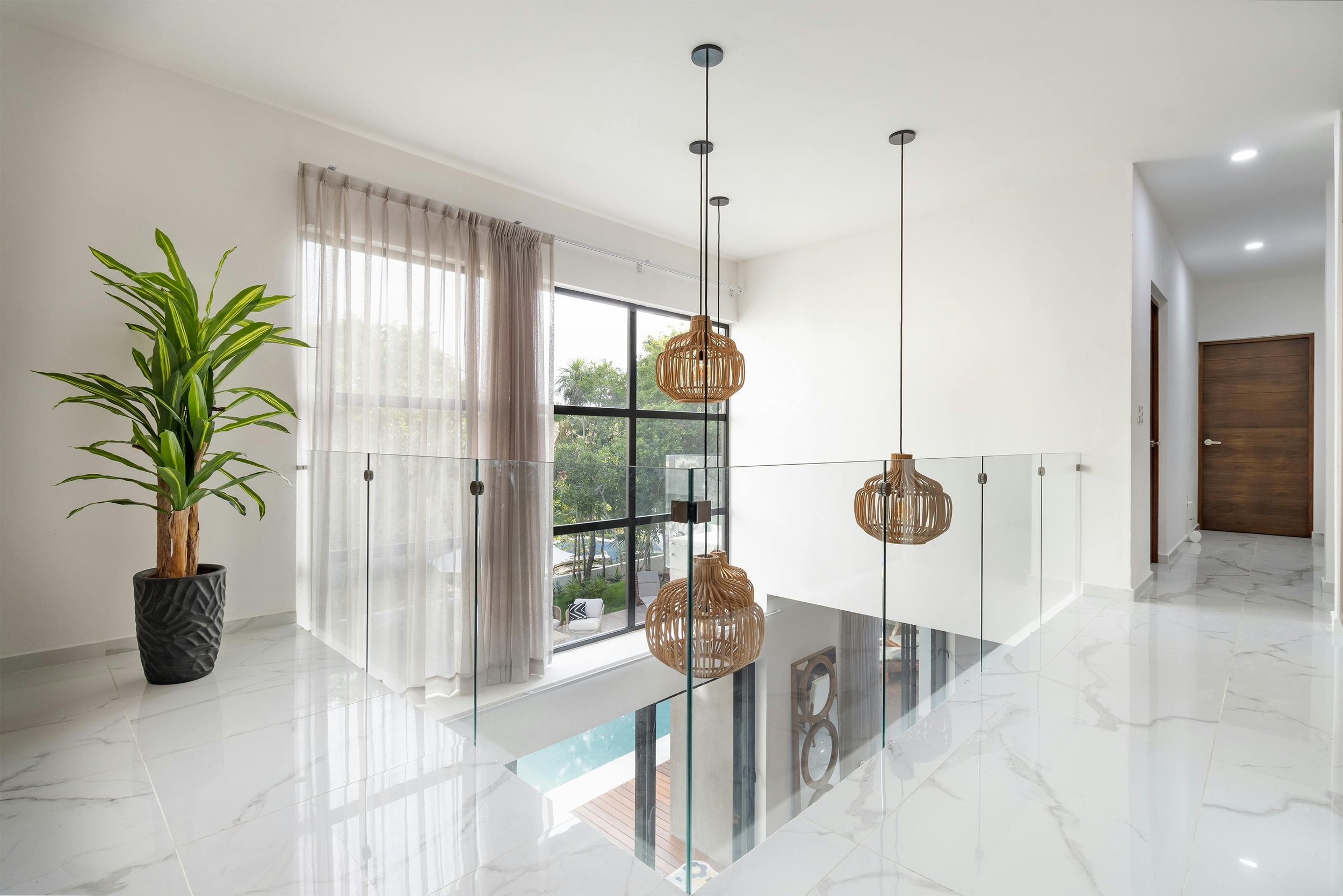 Modern interior hallway with marble floors, glass railing, and wicker pendant lights. A potted plant is next to a large window with sheer curtains.