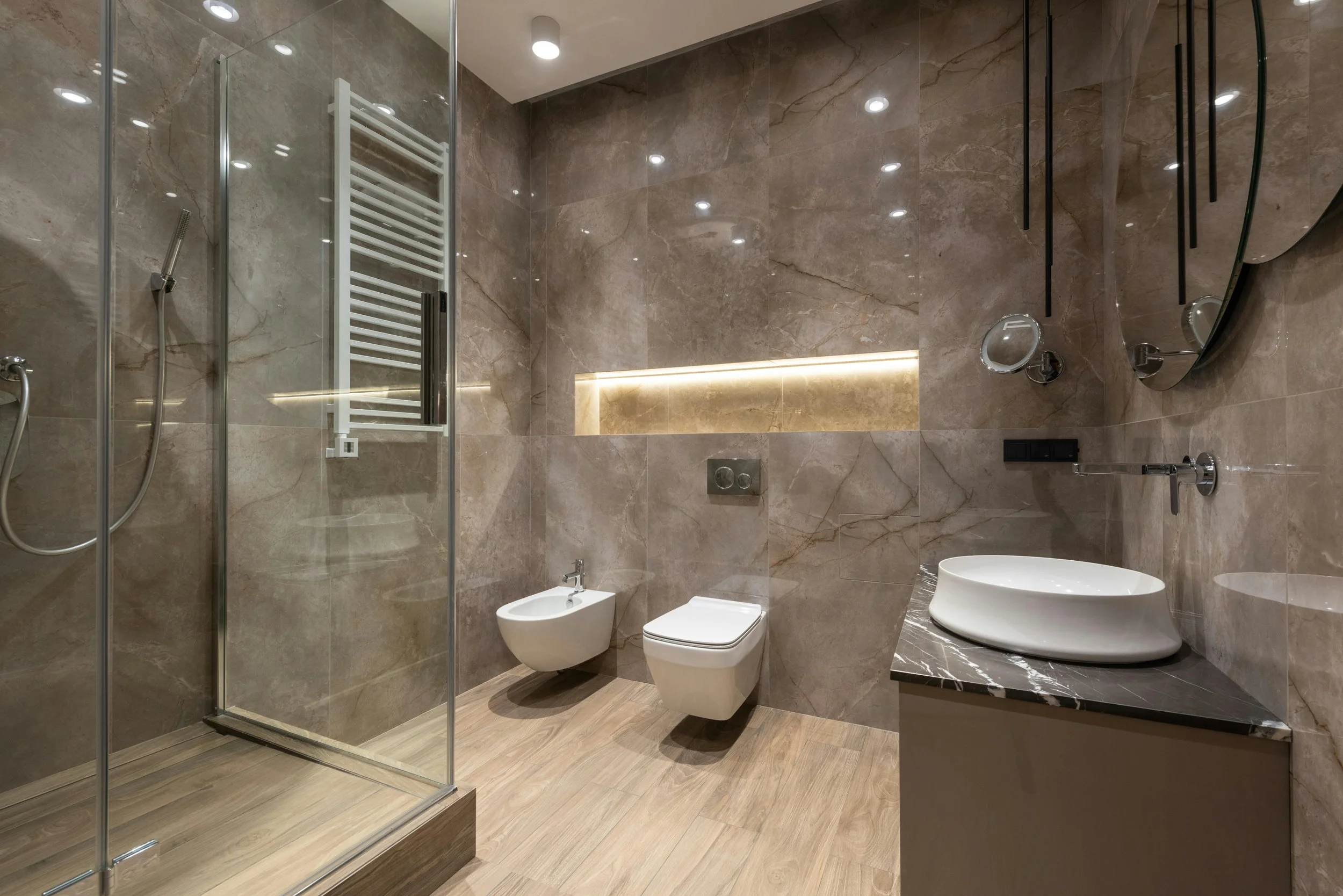 Modern bathroom with glass shower, wall-mounted toilet and bidet, round mirrors, vessel sink, and stone tiles.