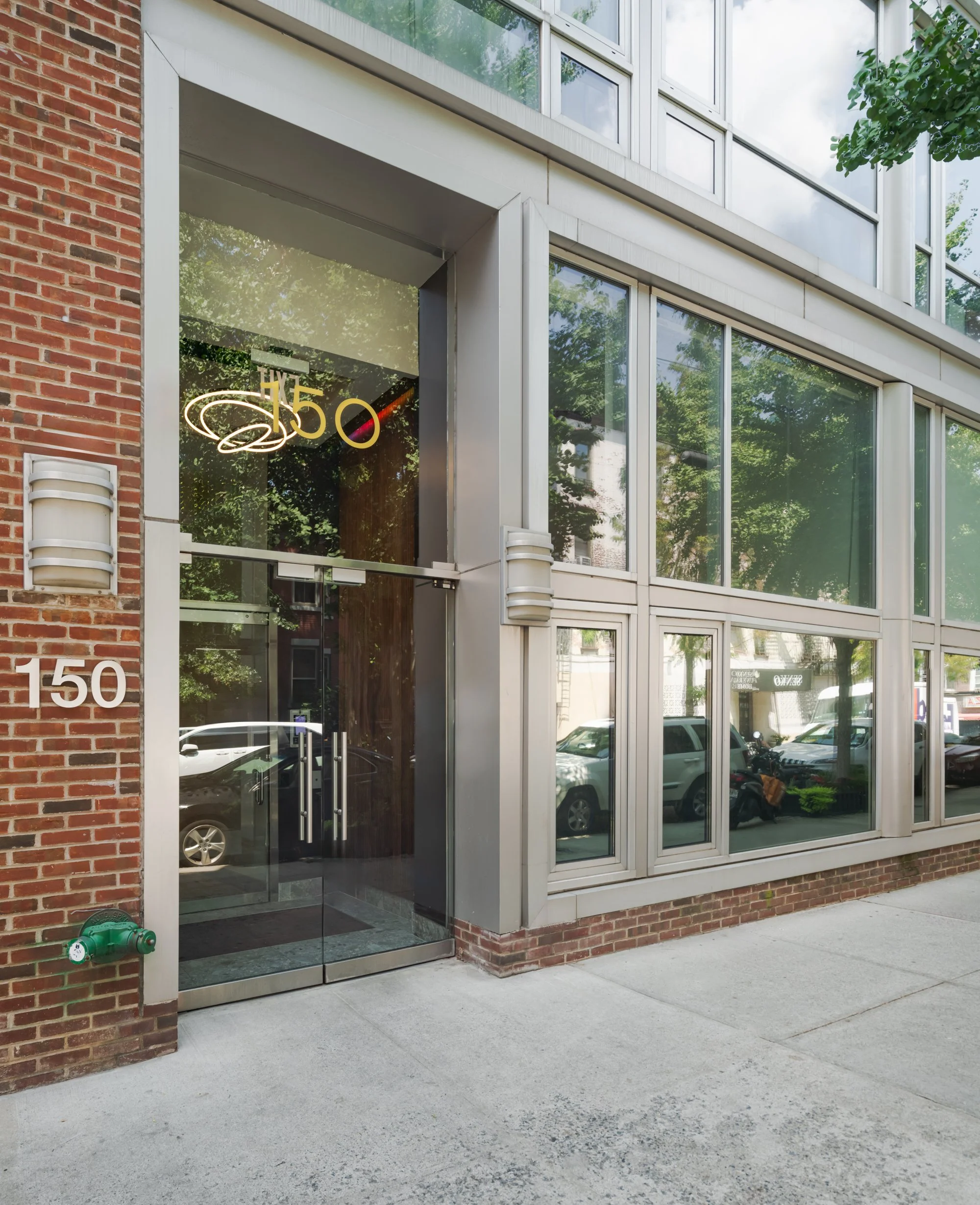 Modern building entrance with glass doors and large glass windows, bearing the number 150, and a neon sign inside that reads 'H5' with decorative elements.