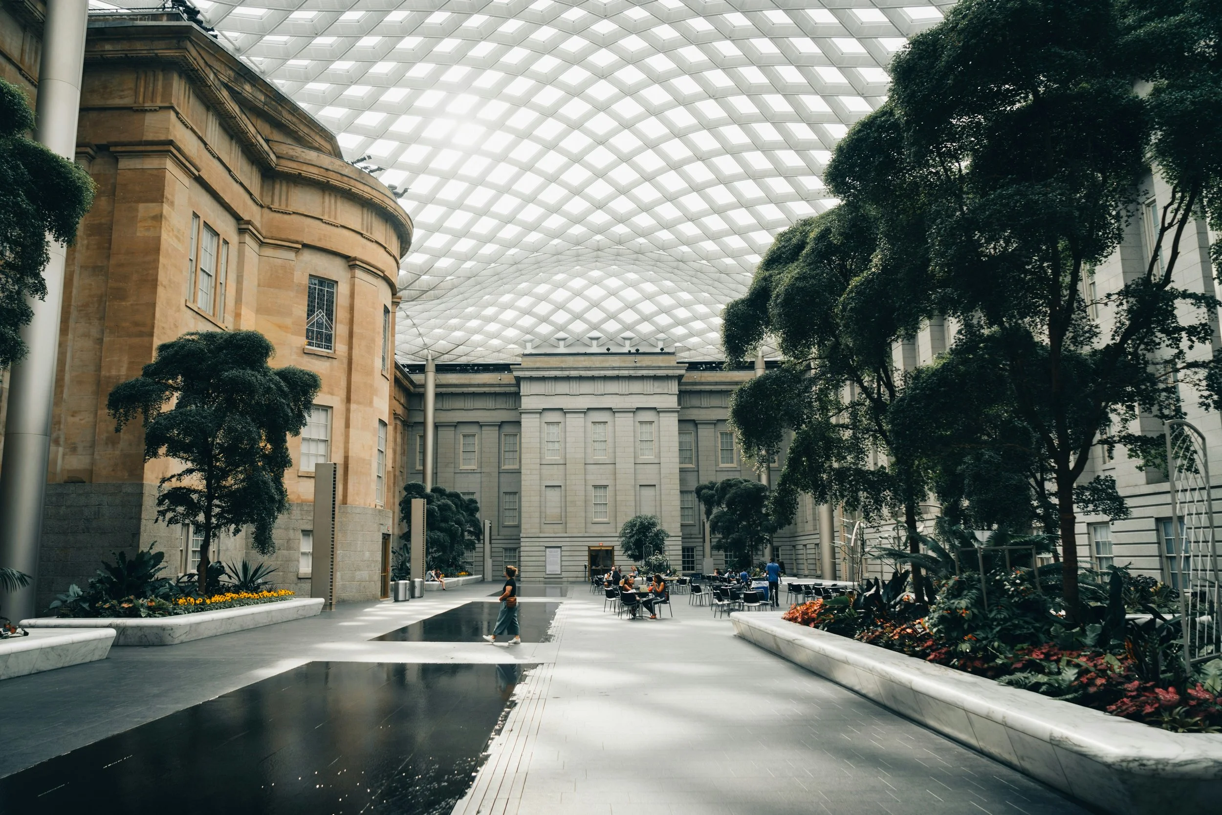 Interior of a large atrium with a glass roof, featuring classical architecture, lush greenery, and people sitting at tables.