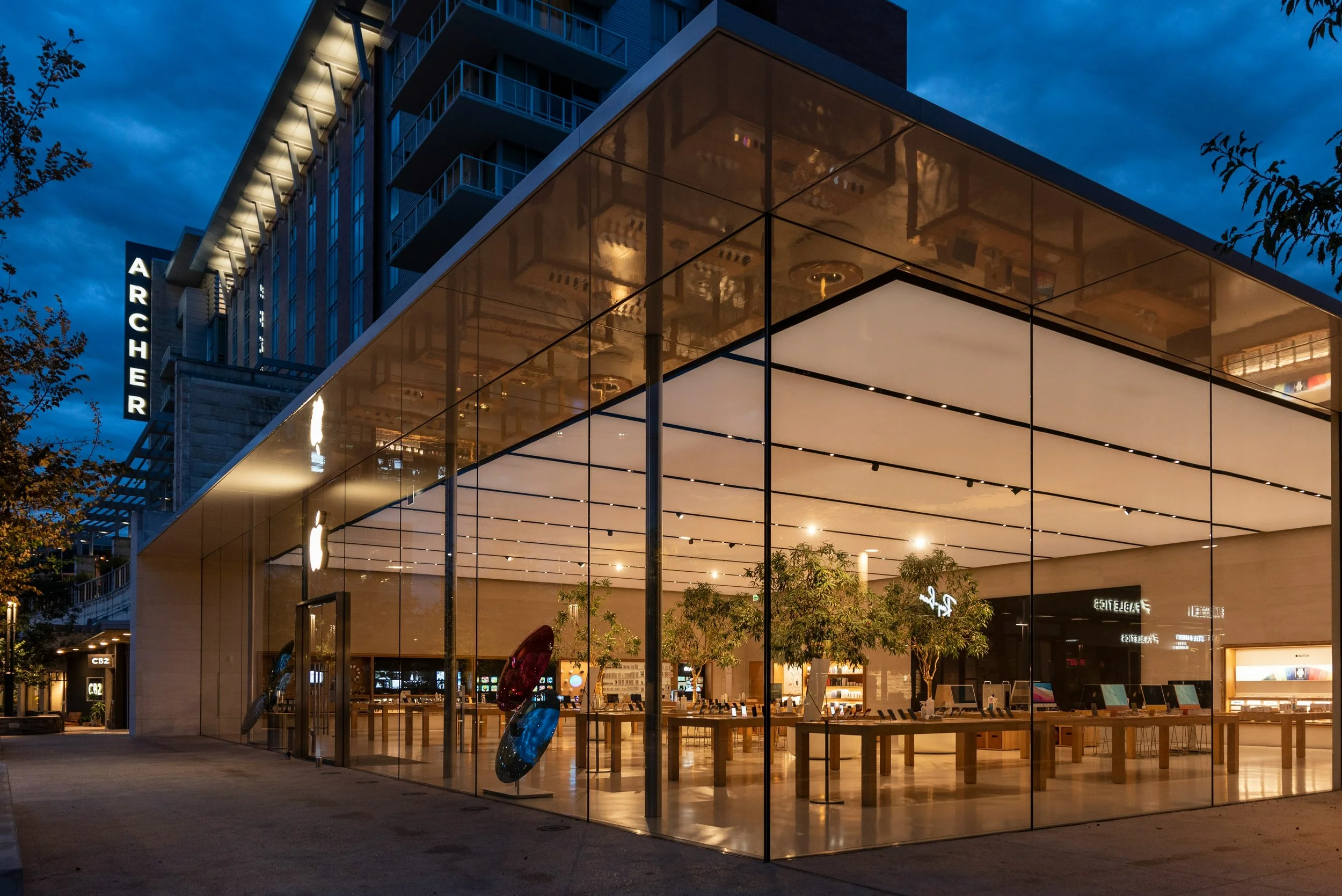Exterior of a modern, glass-fronted store at night, showcasing illuminated interior with trees and electronic products displayed on tables. Nearby building has 'Archer' sign illuminated.