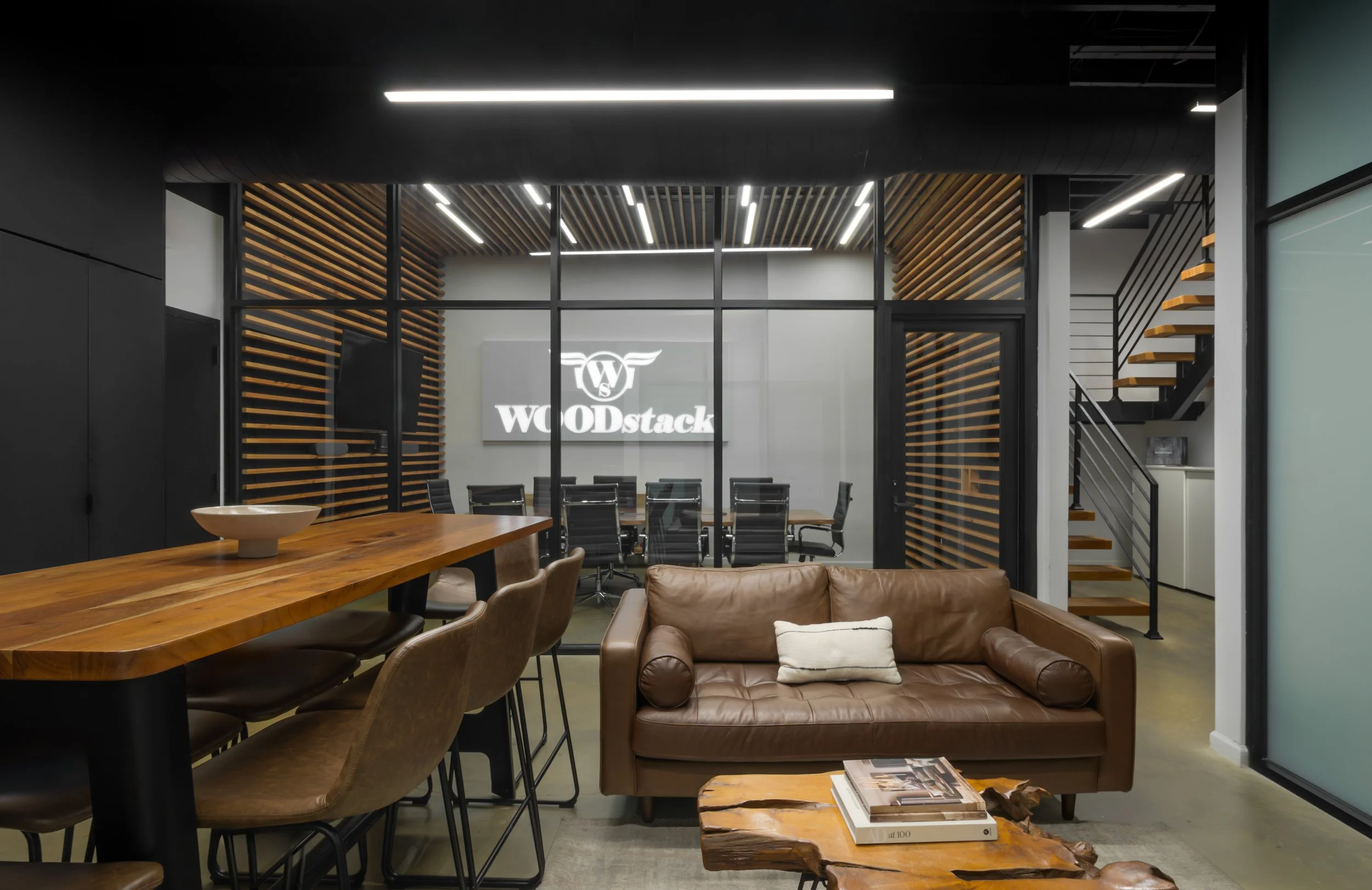 Modern office lounge with brown leather sofa, wooden coffee table, high counter with brown chairs, and glass-walled conference room with logo "WOODstack" in the background.