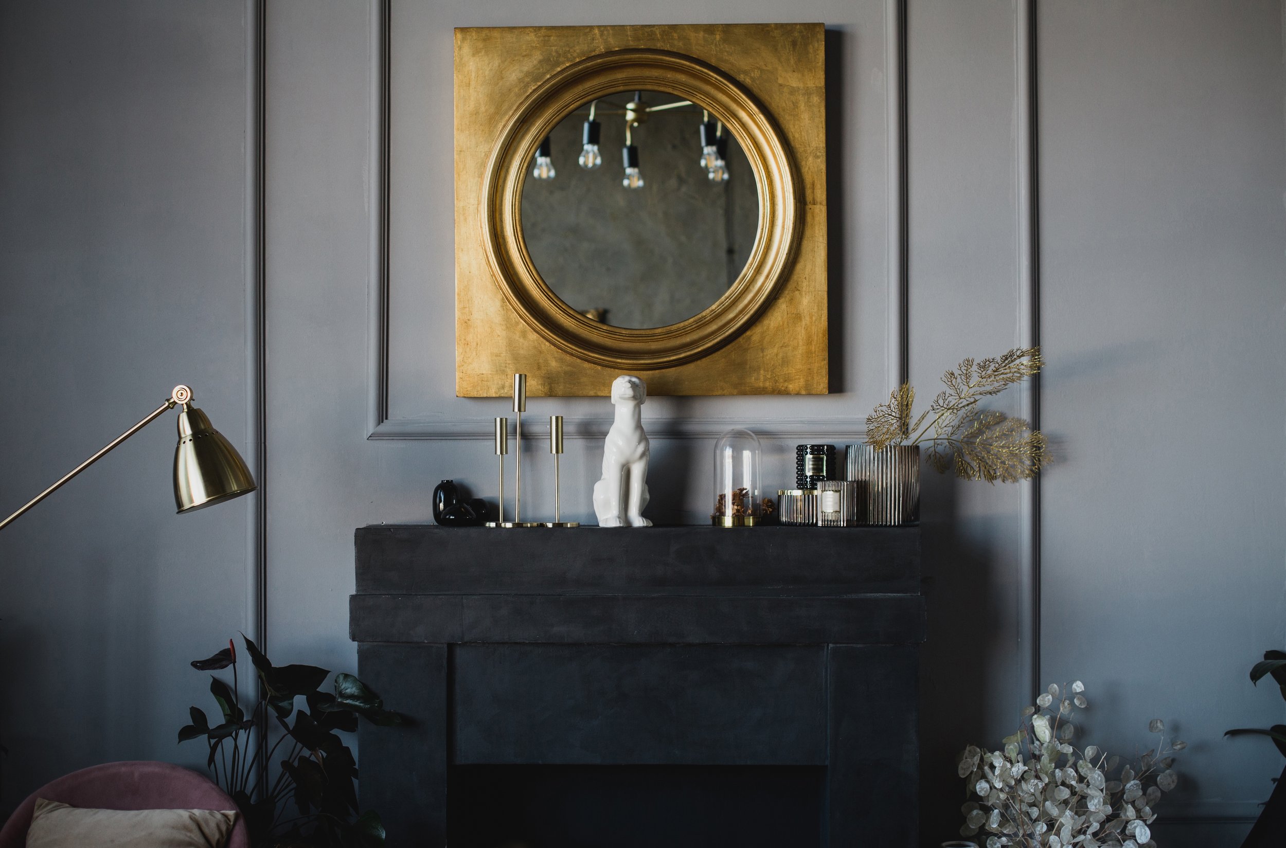 Elegant interior with gray walls, a gold-framed circular mirror, a dark-toned fireplace mantel with decor including a white dog figurine, candles, plants, and a gold floor lamp.