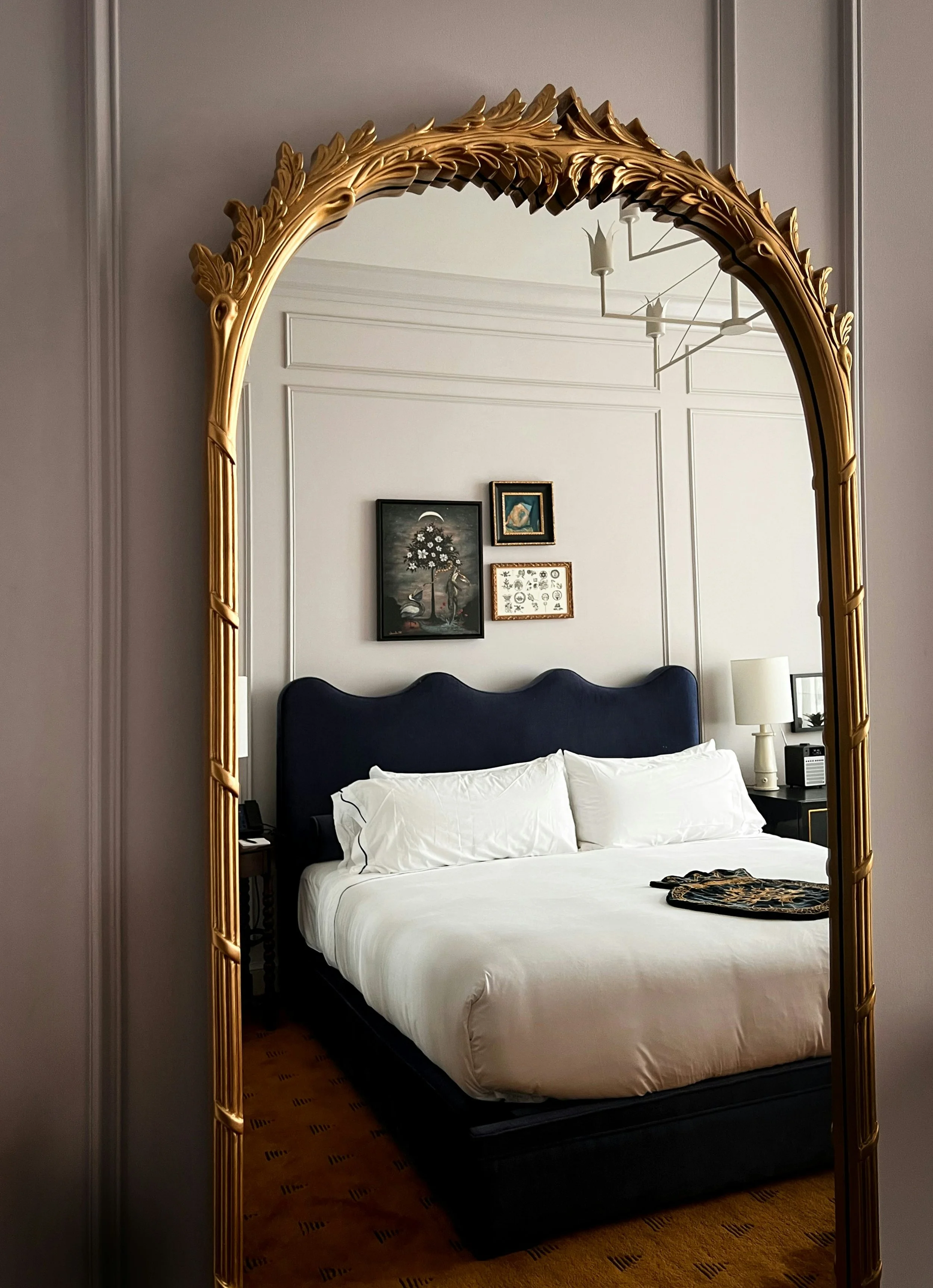 Elegant bedroom reflected in ornate gold mirror, featuring a navy upholstered headboard, crisp white bedding, framed artwork on the wall, and a side table with a lamp.