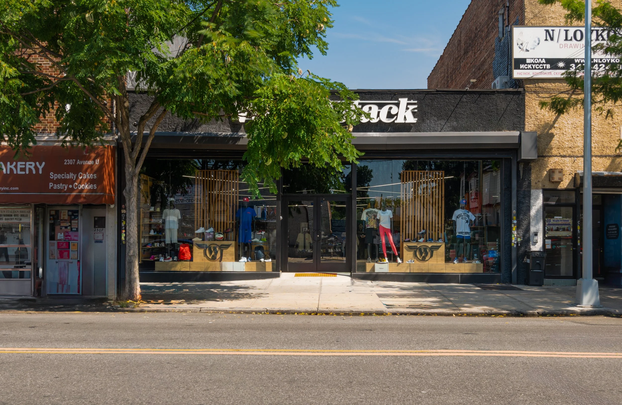 Storefront with mannequins displaying clothing, basketballs, and sports accessories inside. A large tree with green leaves is in front, partially obscuring the view. The store has a black awning with white lettering and is situated between a bakery and another business on a city street.