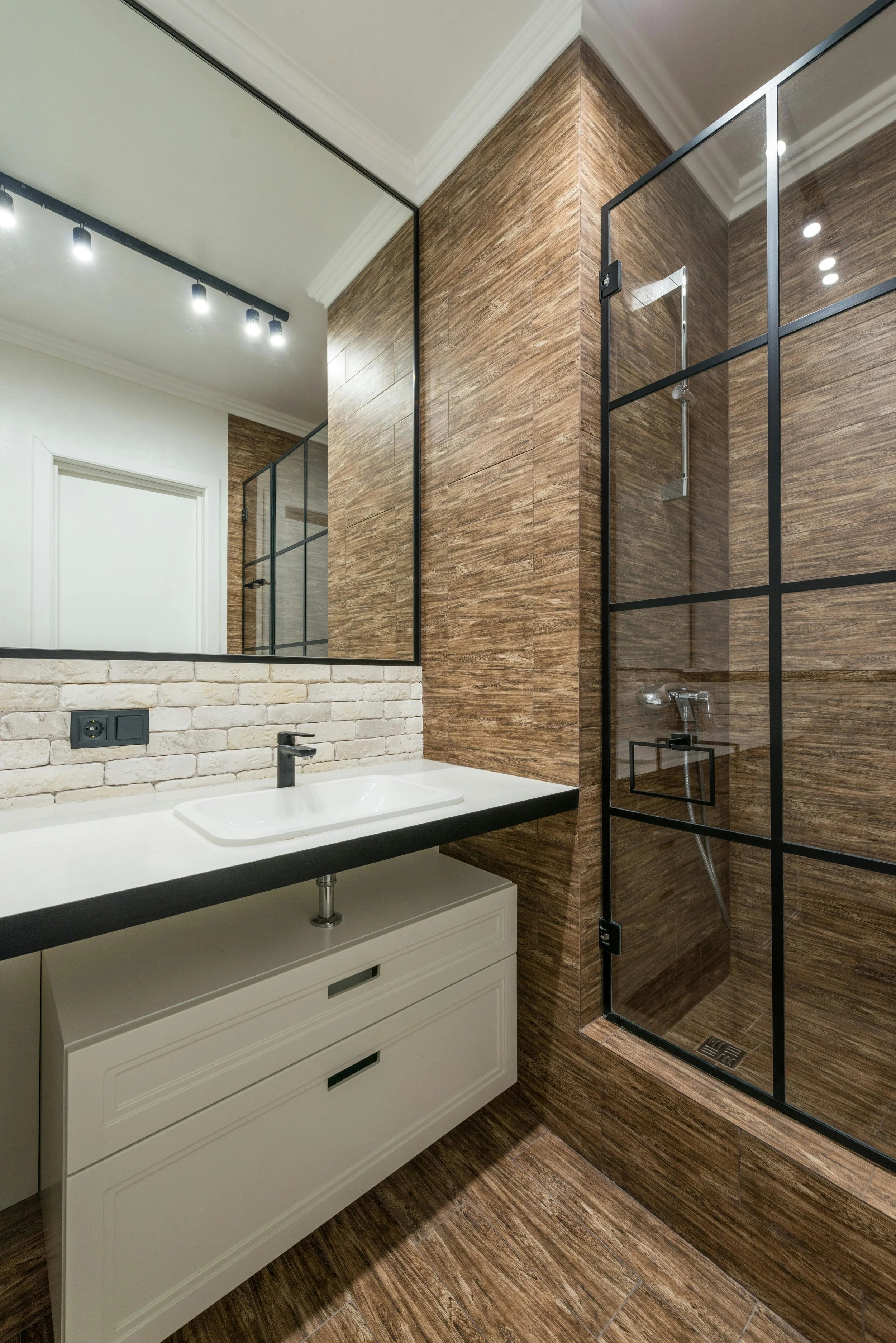 Modern bathroom with a white sink, large mirror, wood-textured tiles, and glass-enclosed shower.