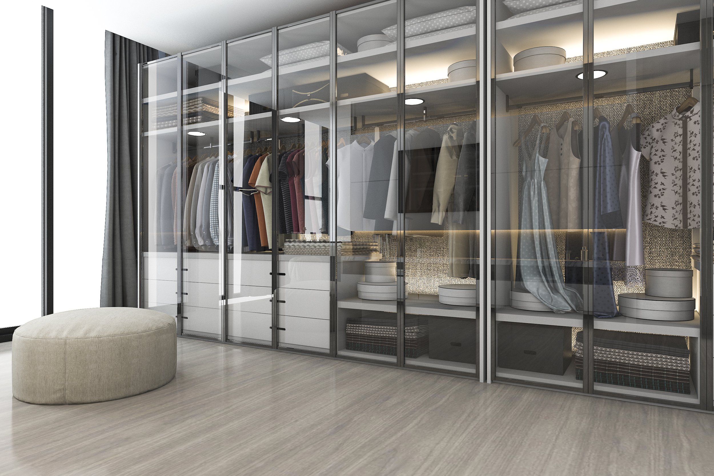 Modern walk-in closet with glass doors, neatly organized clothing, shelves with storage boxes, and a beige pouf on a wooden floor.