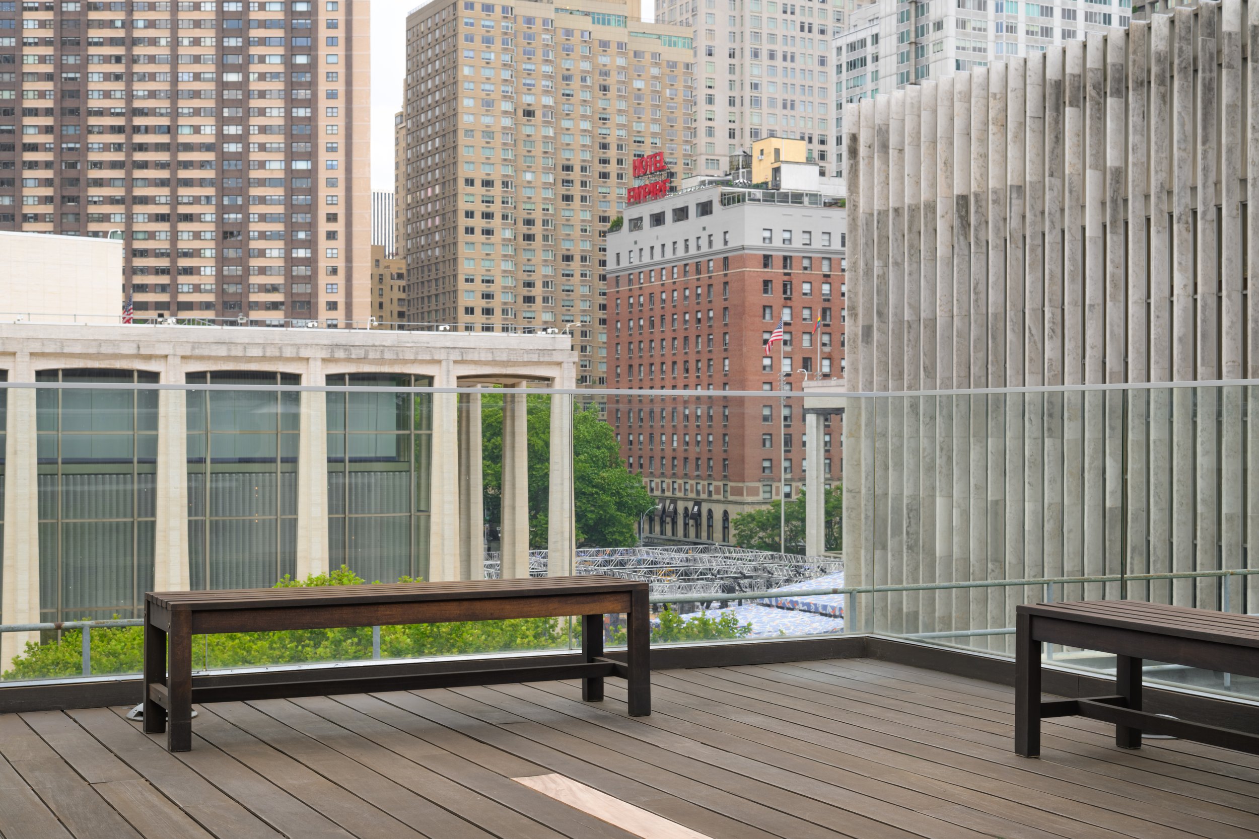 Urban cityscape viewed from a balcony with wooden flooring, two benches, glass railing, and tall buildings in the background.