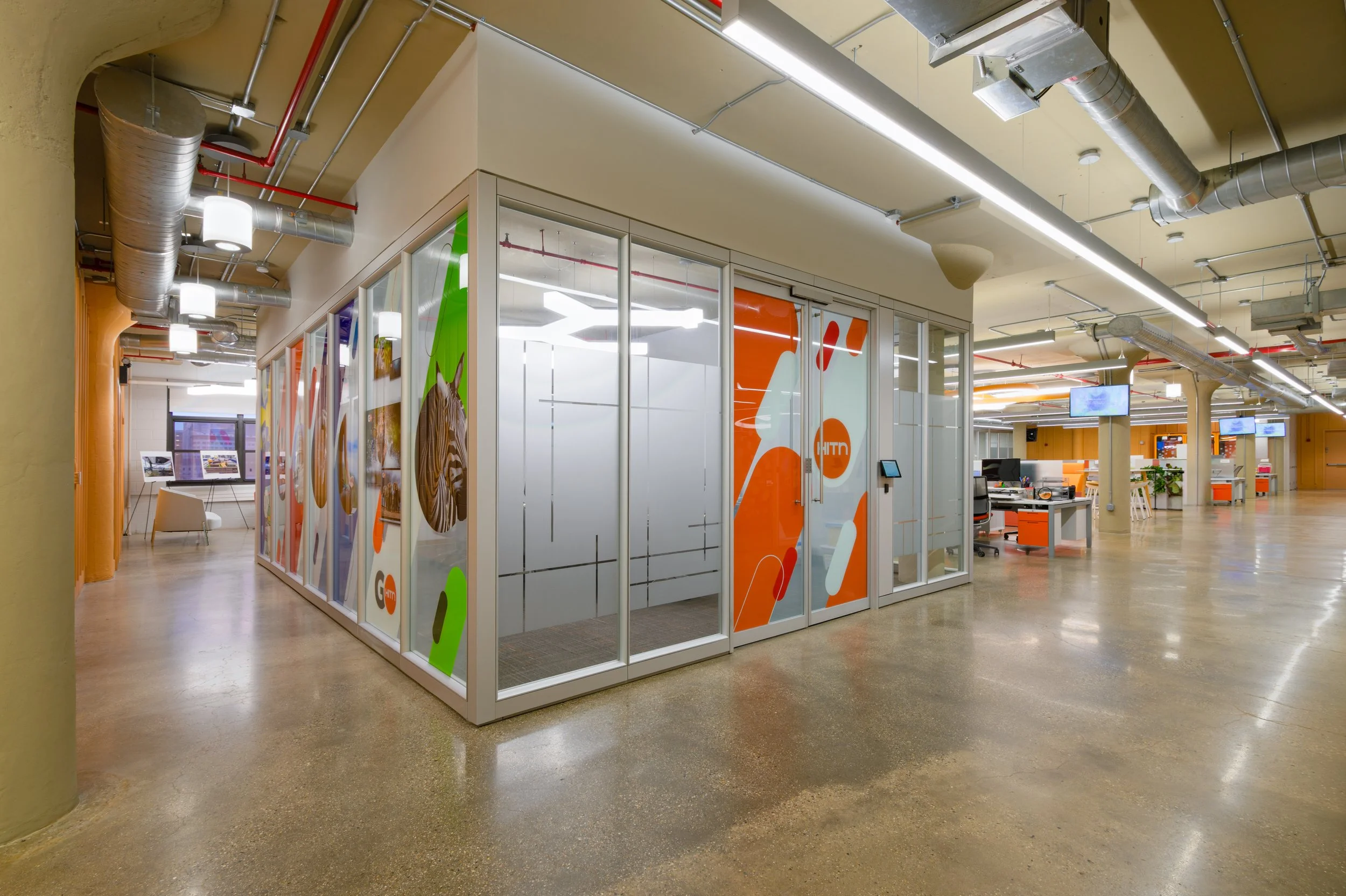 Modern office space with glass enclosed meeting room featuring colorful graphics on the glass, open workstations with computers, and large display screens on the walls. Industrial-style ceiling with exposed ductwork and lighting.