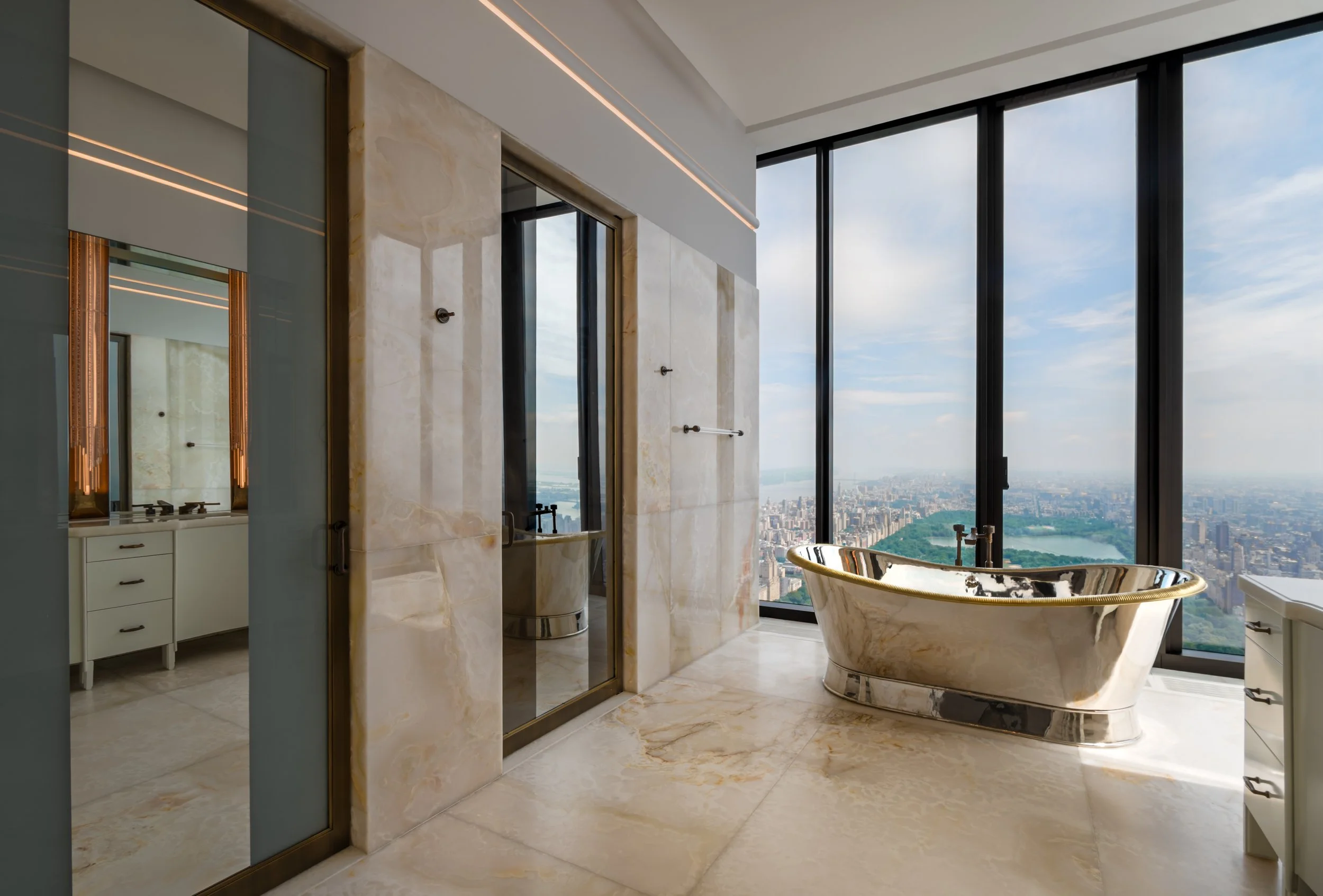 Luxurious bathroom with a freestanding bathtub with a gold trim, large windows showing city skyline and central park, marble walls and floor, and a separate shower area.