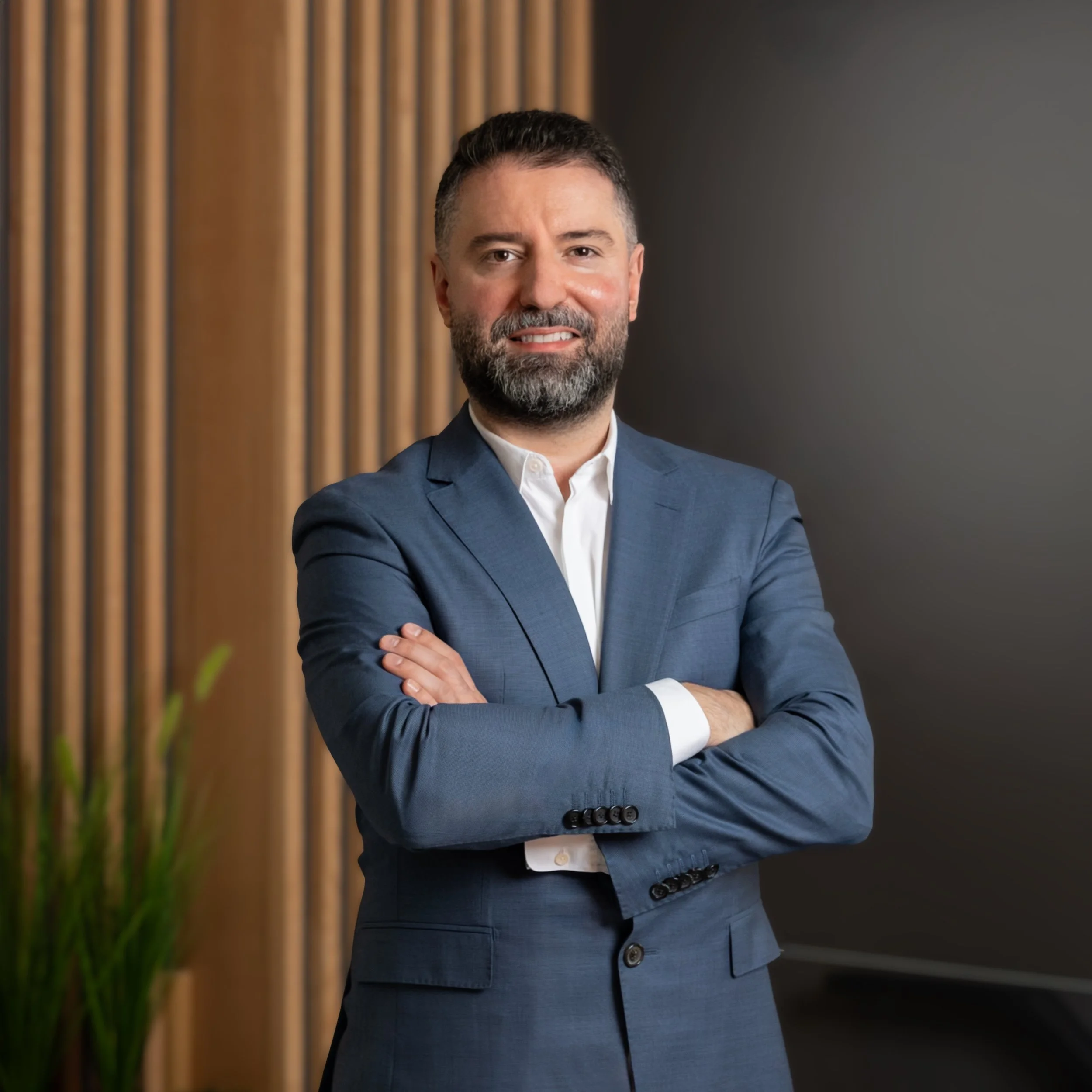 A man with a beard wearing a white shirt and dark blazer stands in front of a wooden slatted background, with plant decor on the side.
