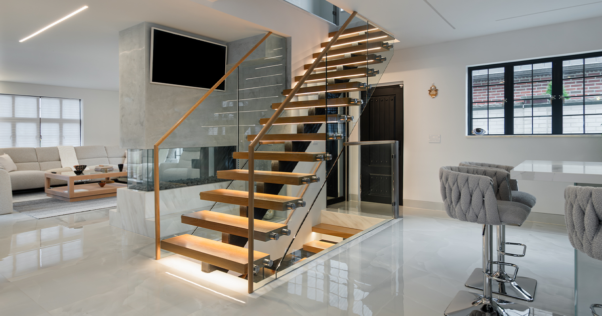 Modern living room with a floating staircase, beige sectional sofa, and flat-screen TV, featuring bright lighting and sleek decor.