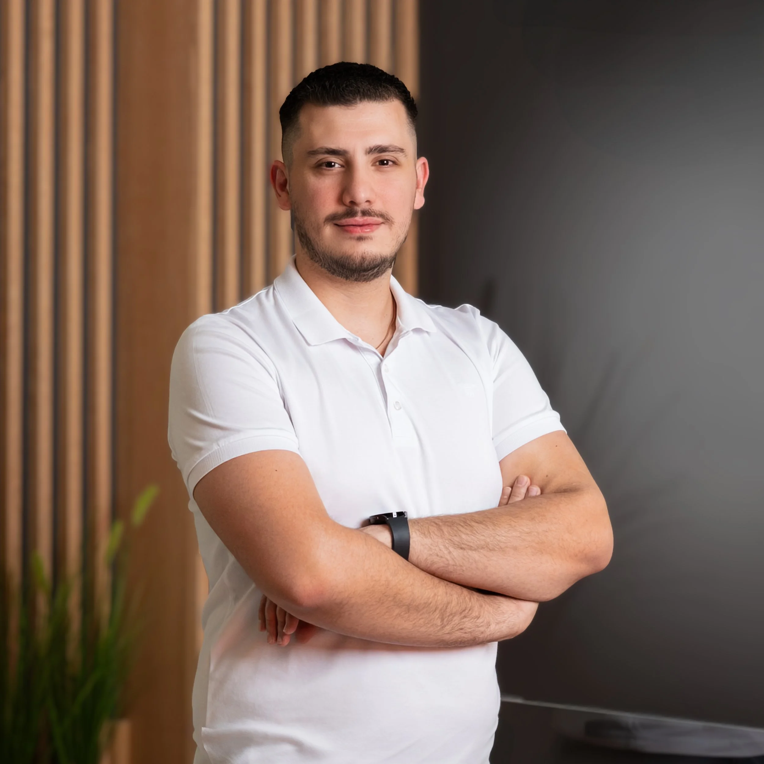 A young man with short dark hair and a beard, wearing a white polo shirt and a black smartwatch, standing indoors with arms crossed. Background features a wooden wall and a dark wall with a potted plant visible in the corner.