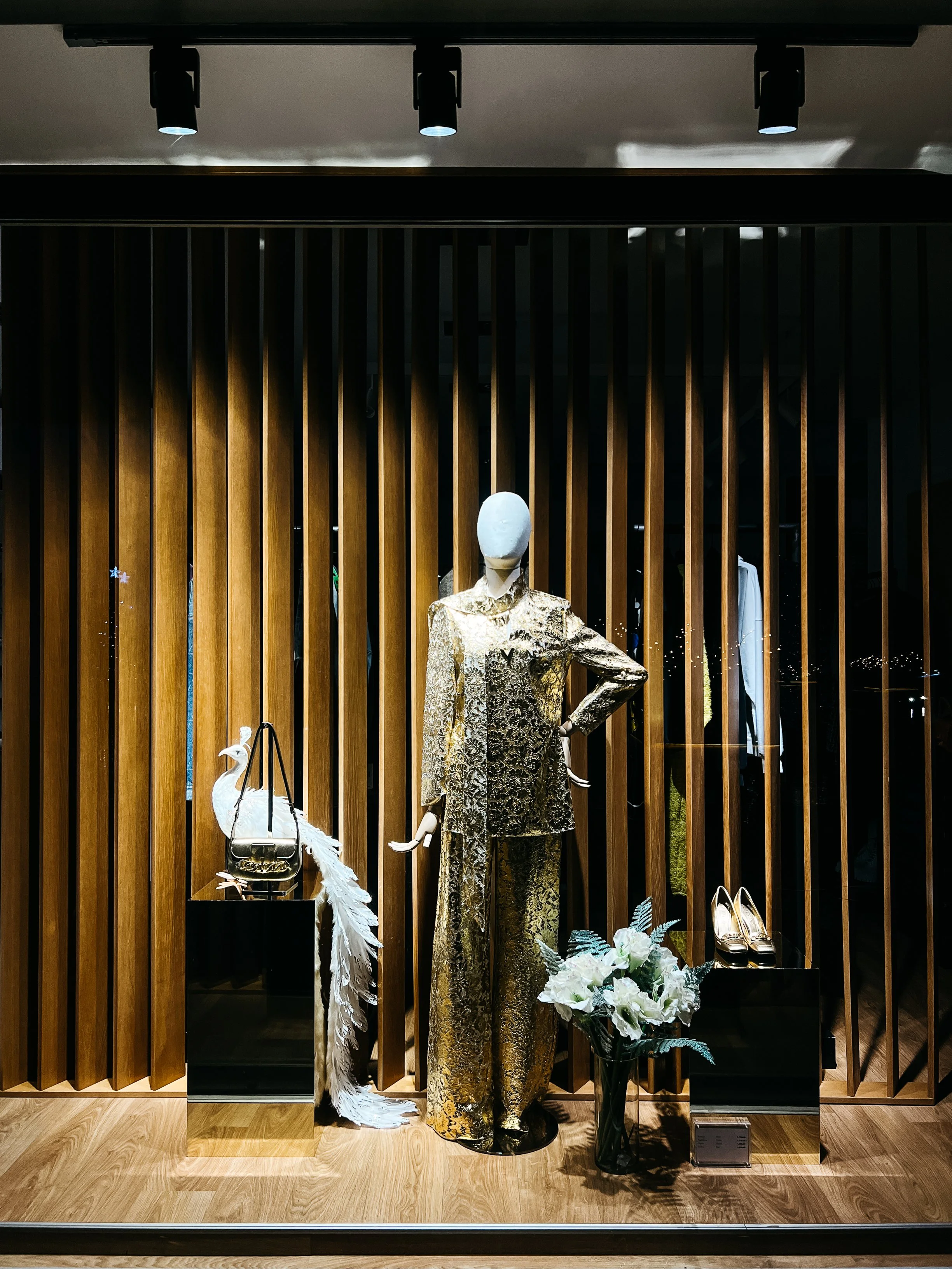 Storefront display featuring a mannequin dressed in a gold, ornate outfit. Beside it is a decorative white peacock figurine on a stand. Gold high heels and a bouquet of white flowers are also displayed. The background features vertical wooden slats and spotlights illuminate the scene.