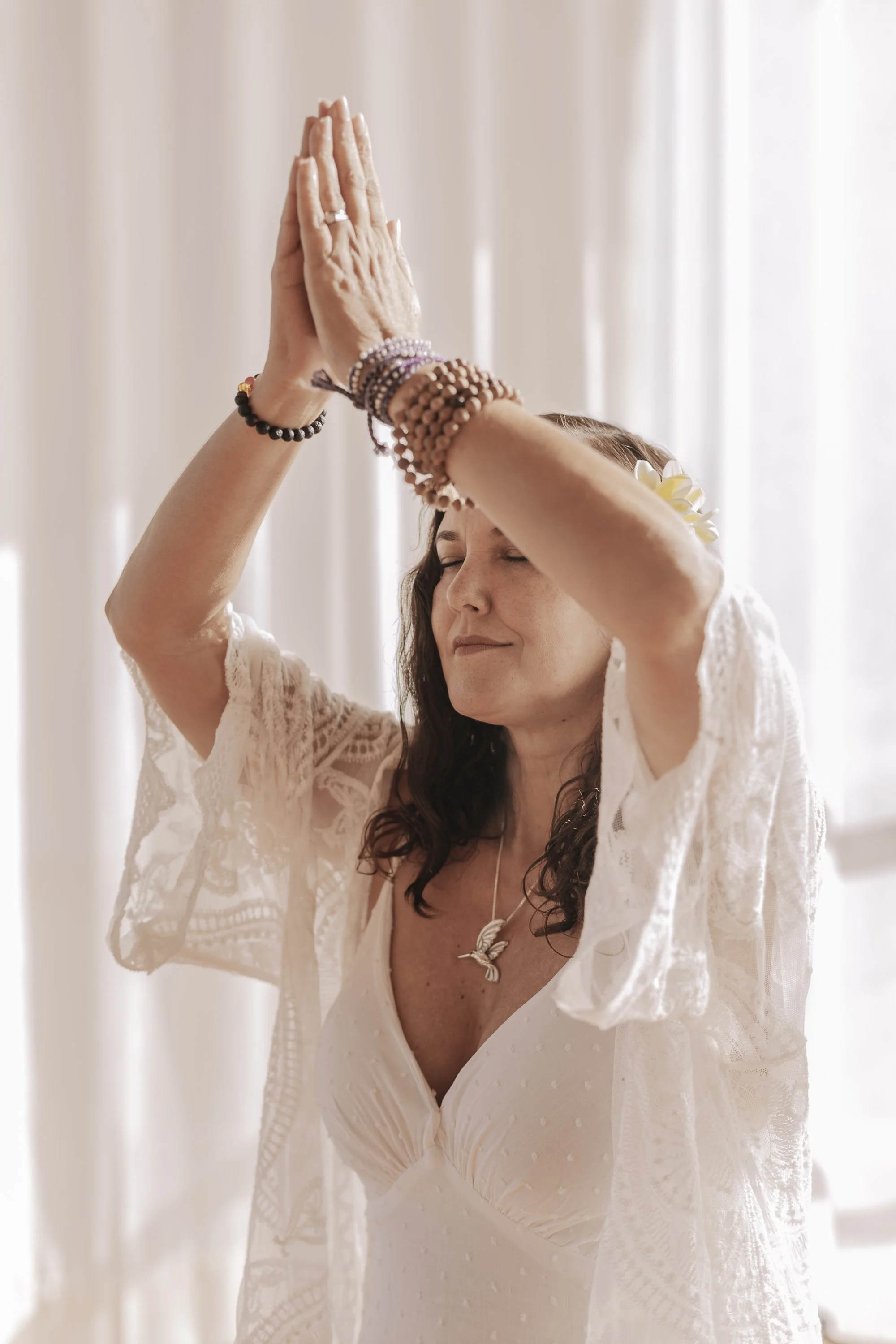 A woman with dark hair, wearing a white dress and jewelry, is praying with her hands together above her head, eyes closed, in an indoor setting with light-colored curtains.
