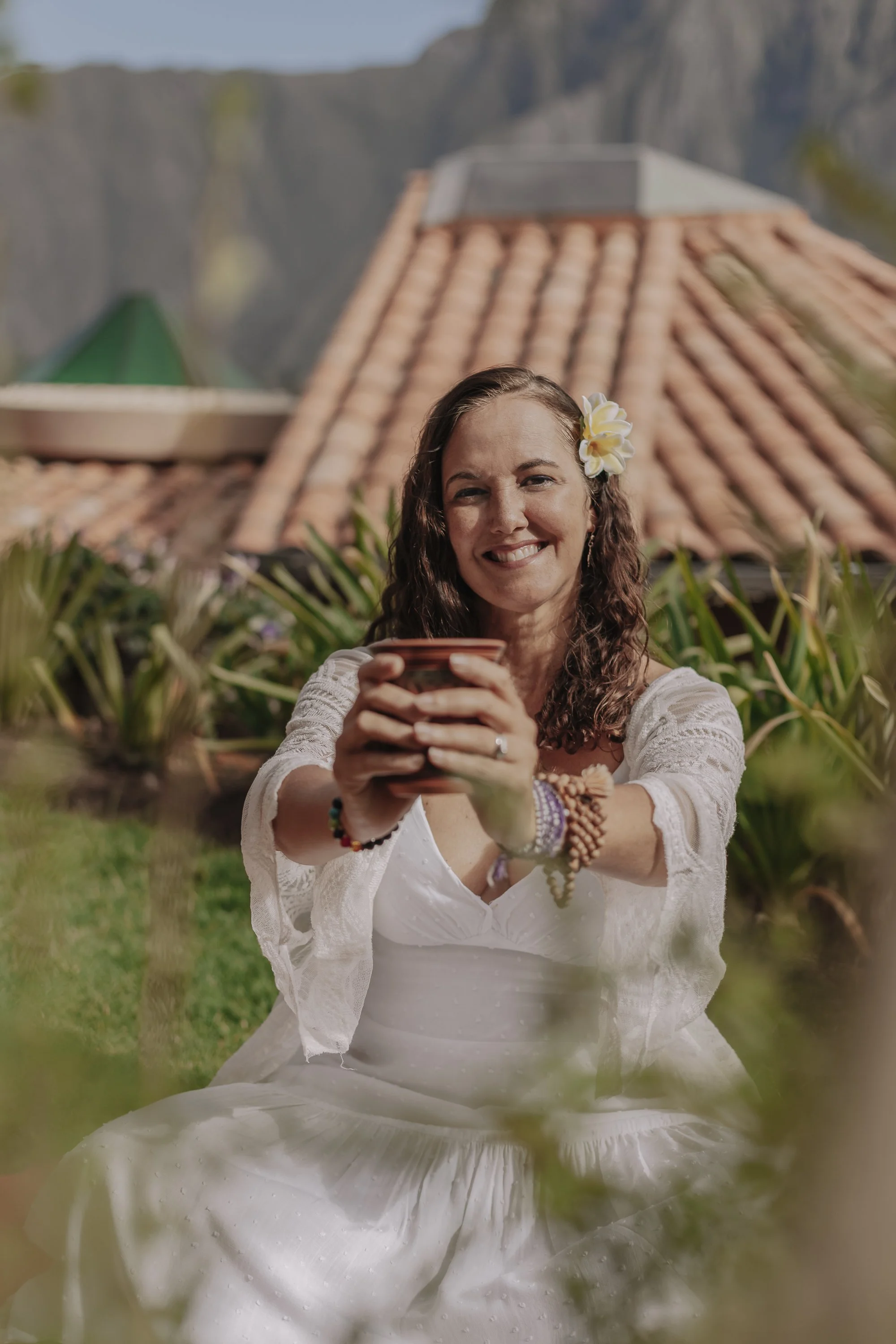 A woman with long curly hair, a flower in her hair, and wearing a white dress and jewelry, smiling and holding a cup outdoors with a red-tiled roof and mountain scenery in the background.