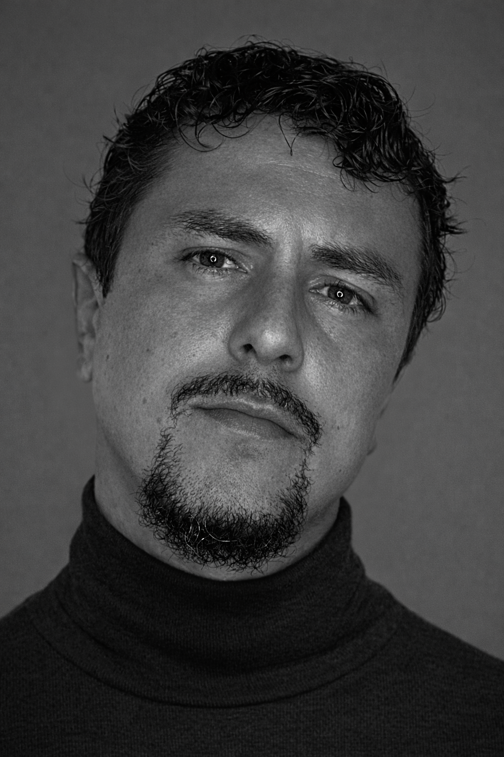 Black and white professional headshot of a man with short curly hair and a goatee, wearing a dark turtleneck and looking confidently at the camera.