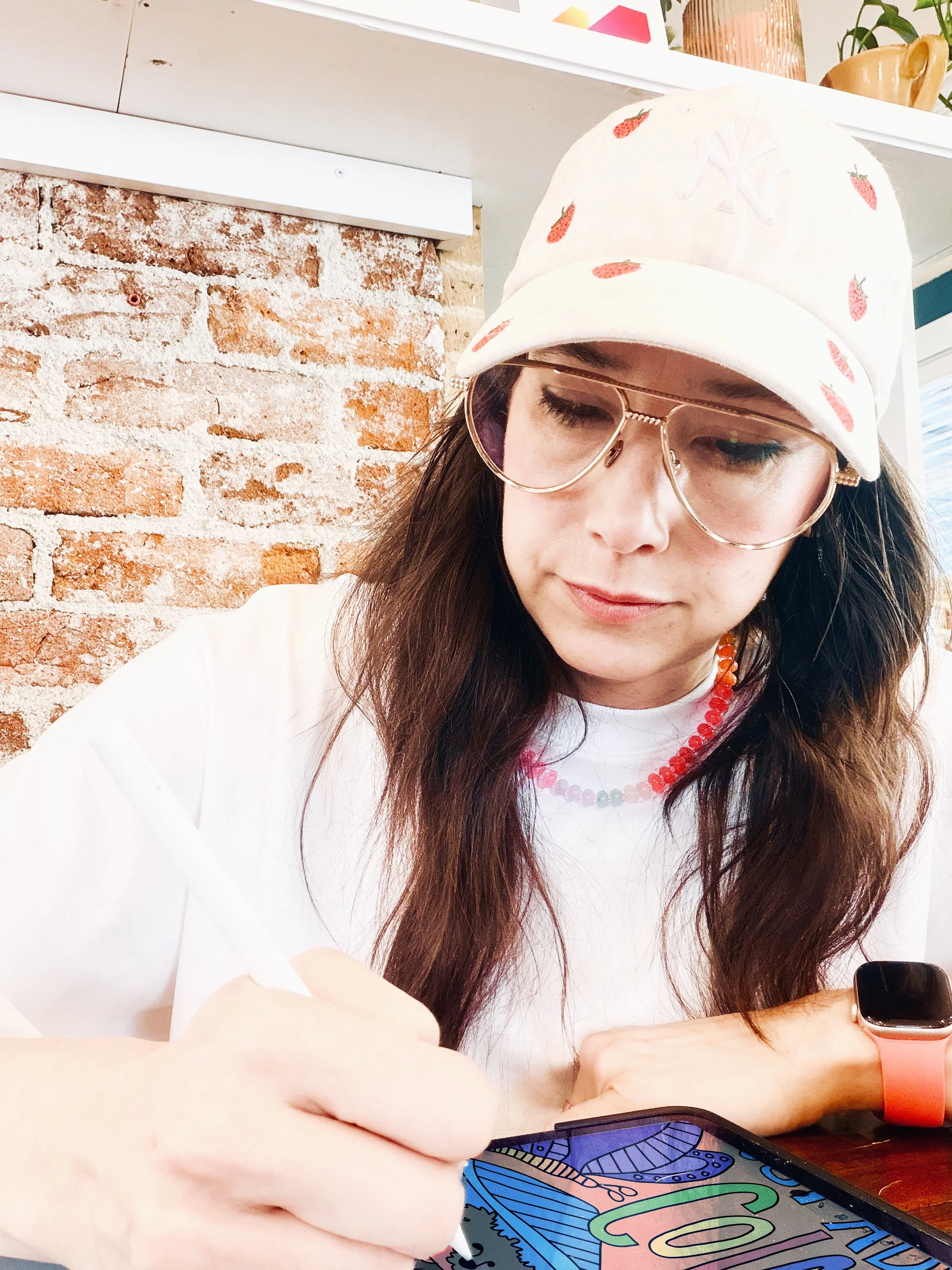Close-up of a young woman wearing a white baseball cap with strawberries, glasses, and a colorful necklace, looking down at her phone with a brick wall and shelf with decorative items in the background.