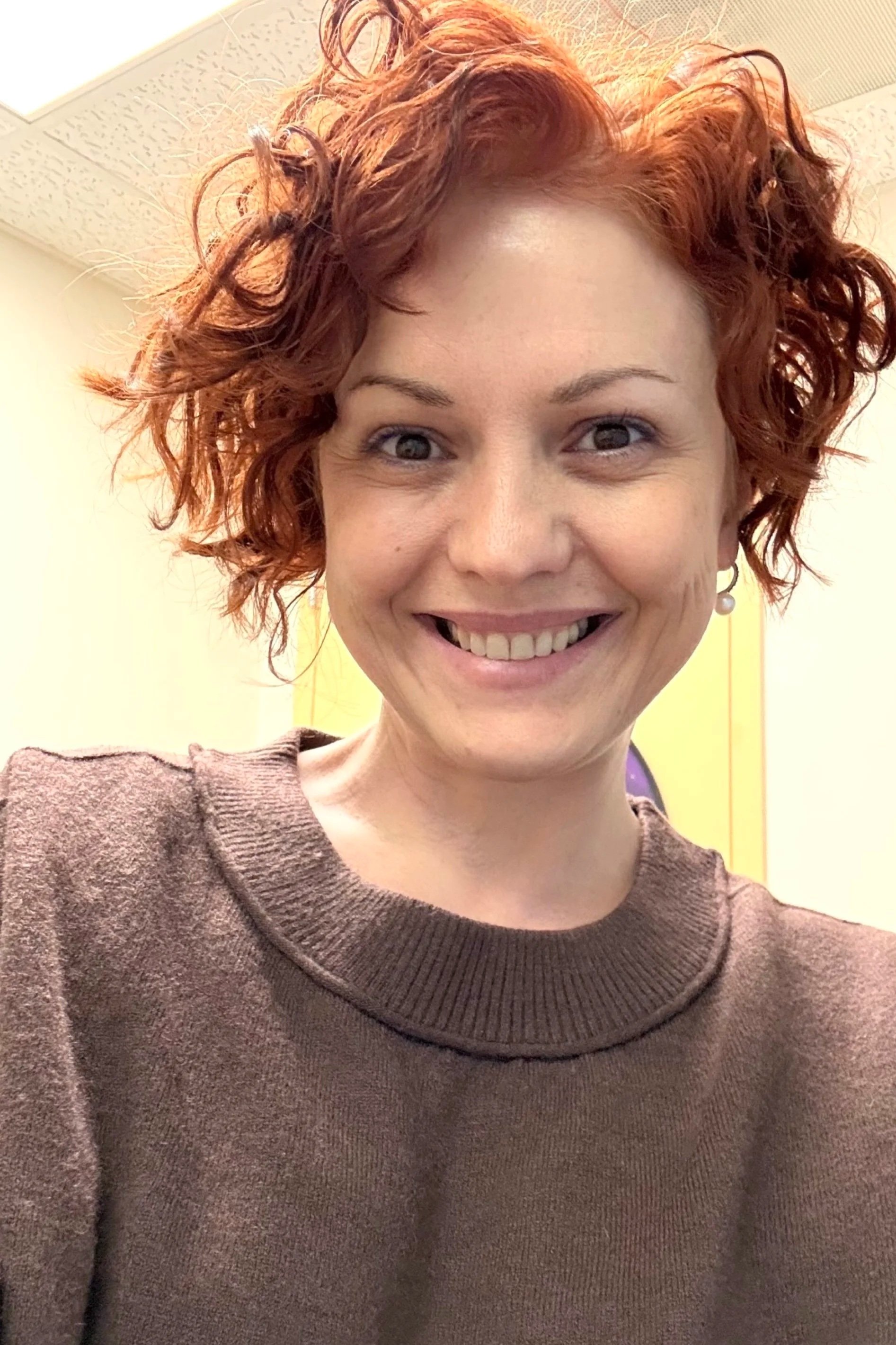 Dana, the owner, sitting on a soft play net in an indoor playroom, smiling and holding onto the mesh sides with a ball pit and climbing wall in the background.