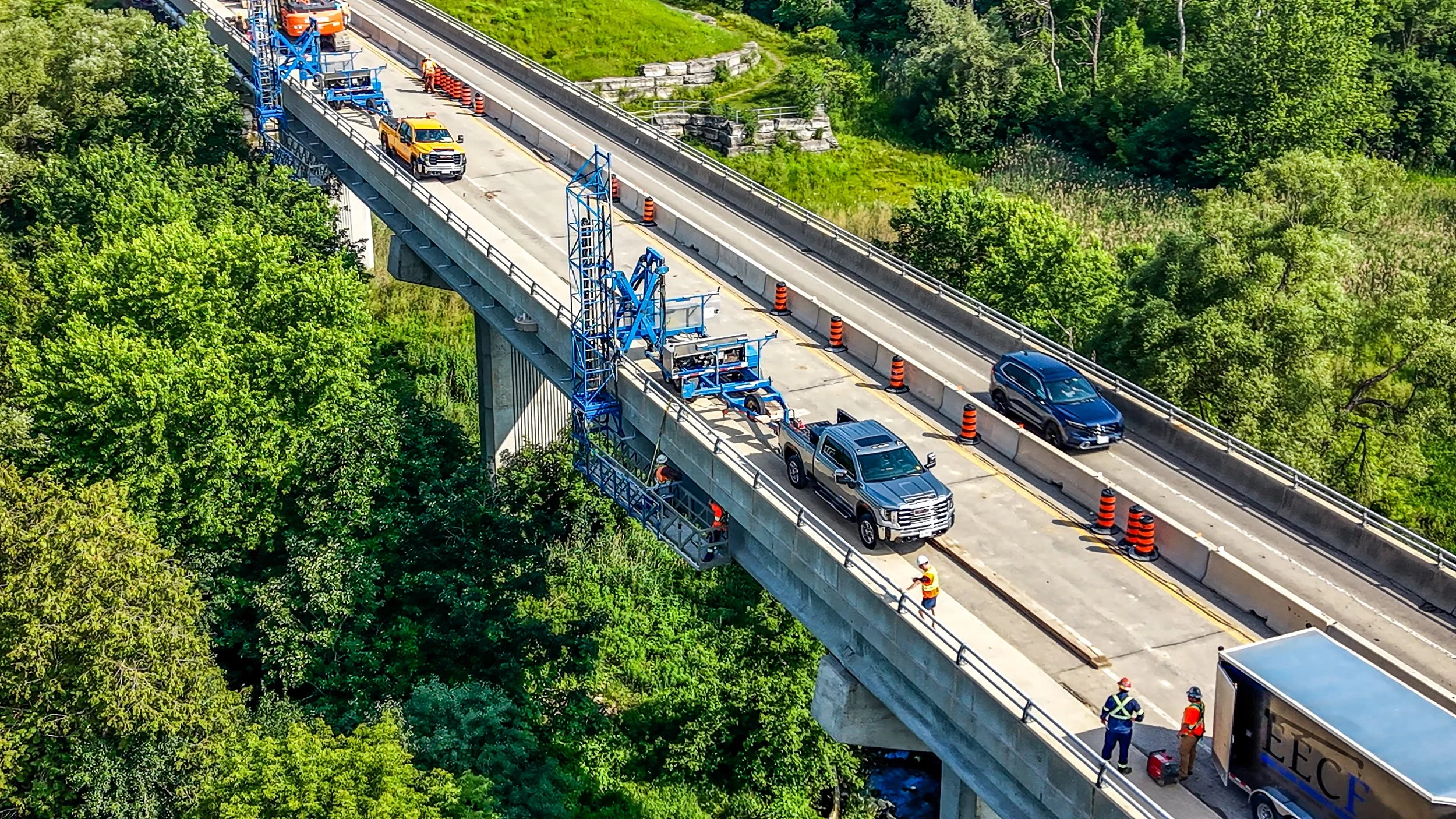Construction workers and vehicles on a bridge undergoing maintenance, with orange cones and equipment present, surrounded by green trees.