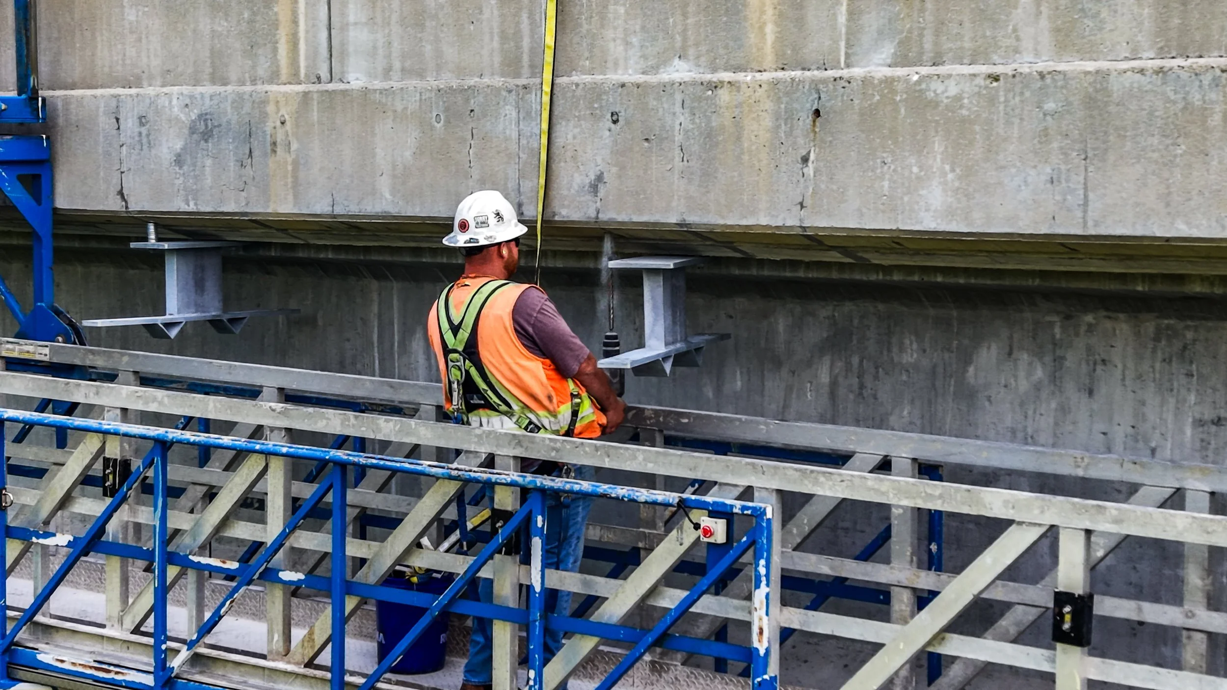 Construction worker wearing a safety helmet and vest, sitting on a scaffold platform at a construction site with concrete walls behind him.