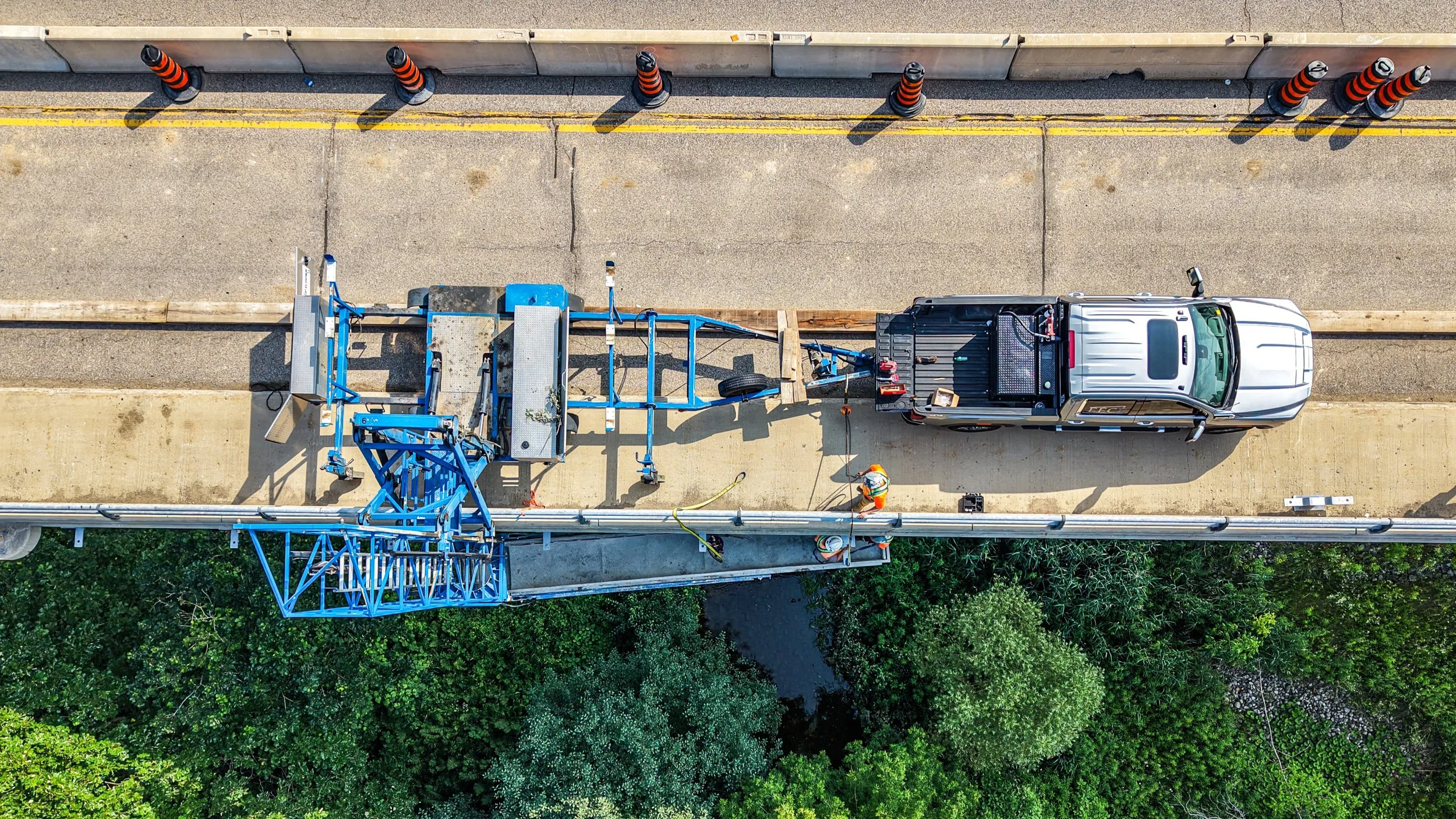 An aerial view of a construction worker operating a blue cherry picker lift on a sidewalk, with orange traffic cones and a concrete barrier, next to a white truck.