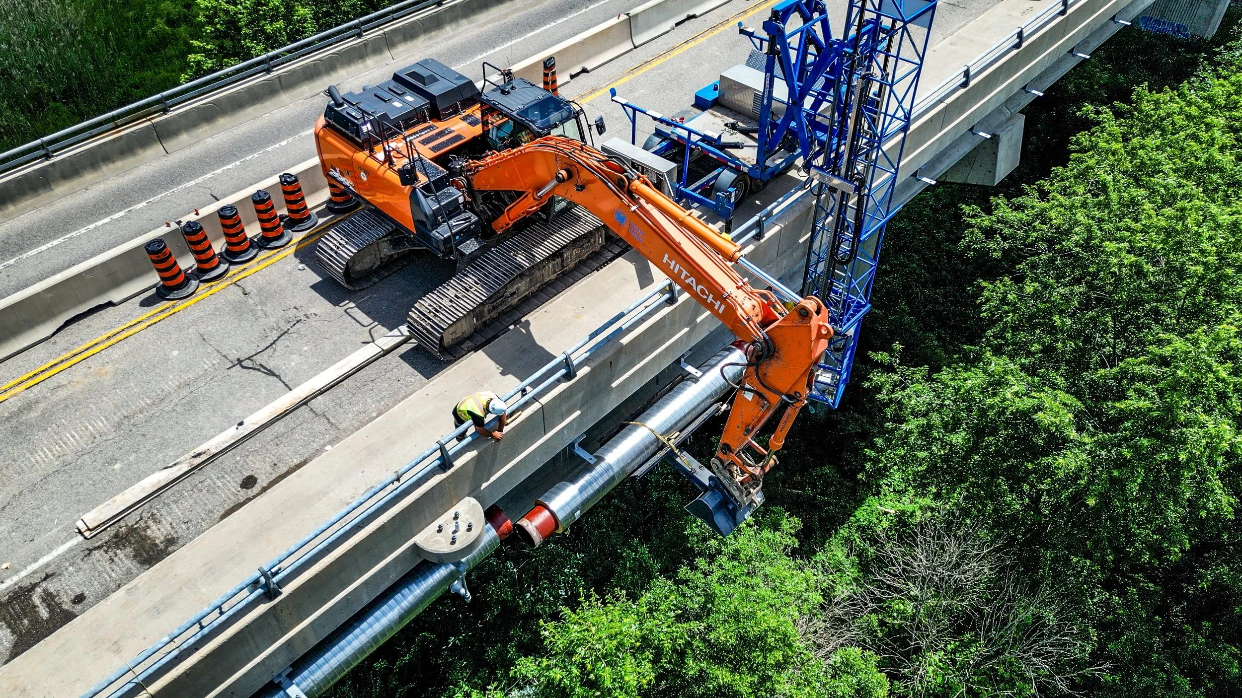 Construction workers using an orange Hitachi excavator to install or repair infrastructure on a bridge or overpass, with traffic cones blocking a lane, alongside green trees.