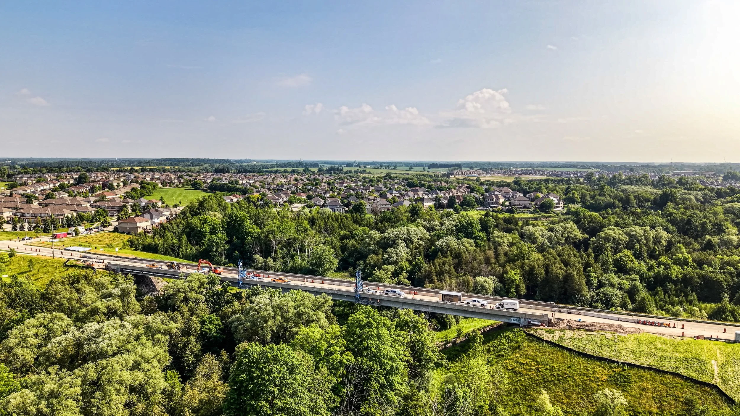 Bird's eye view of a suburban neighborhood with houses, green trees, and a highway under construction, under a partly cloudy sky.