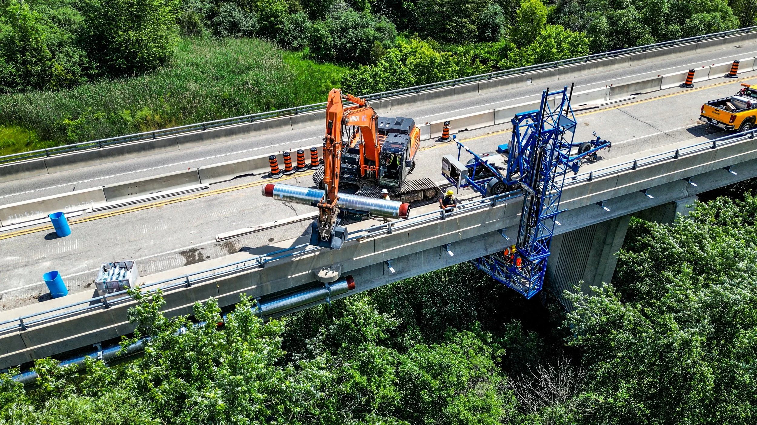 Construction workers and machinery repairing the side of a highway overpass surrounded by green trees.