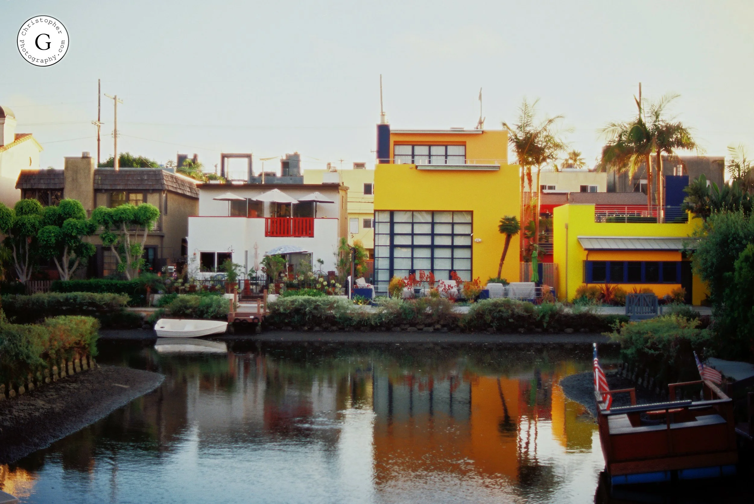 Colorful modern houses along a waterfront with palm trees, reflecting in the water, with a small boat and American flags in the foreground shot on 35mm film .
