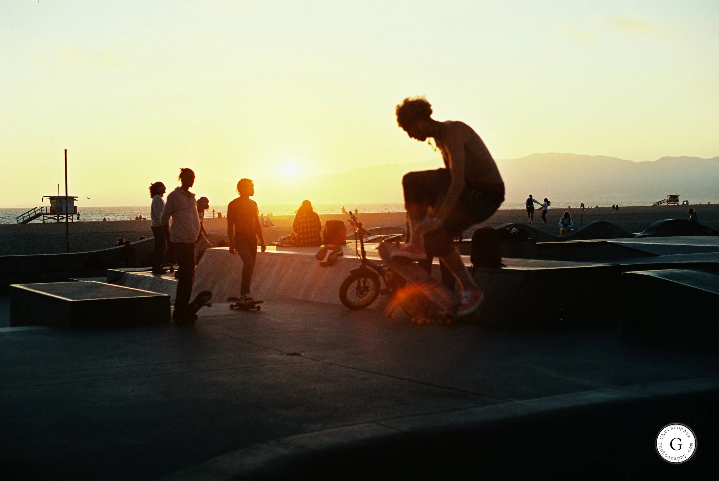 Silhouettes of young people skateboarding and biking at Venice Beach skatepark during sunset shot on 35mm film .