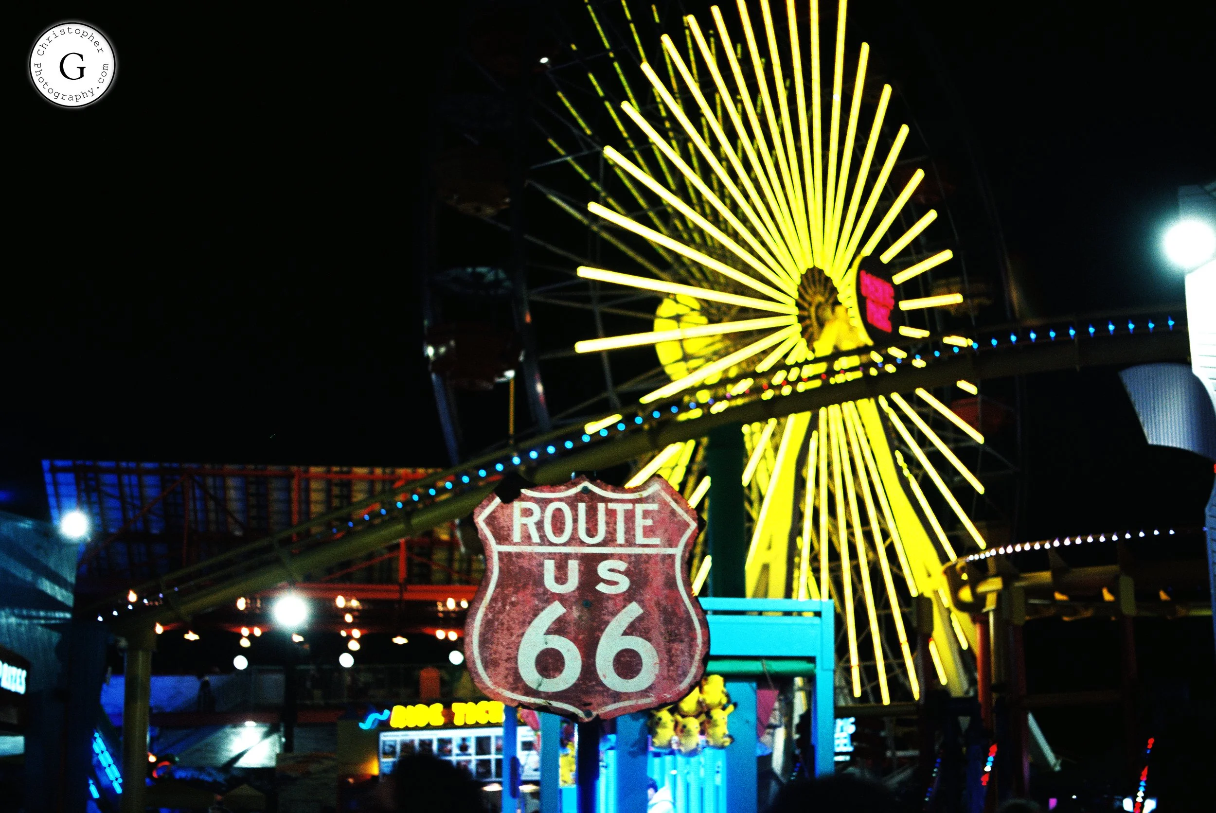 Brightly lit amusement park at night with a large yellow Ferris wheel and a weathered Route 66 sign in the foreground shot on 35mm film.