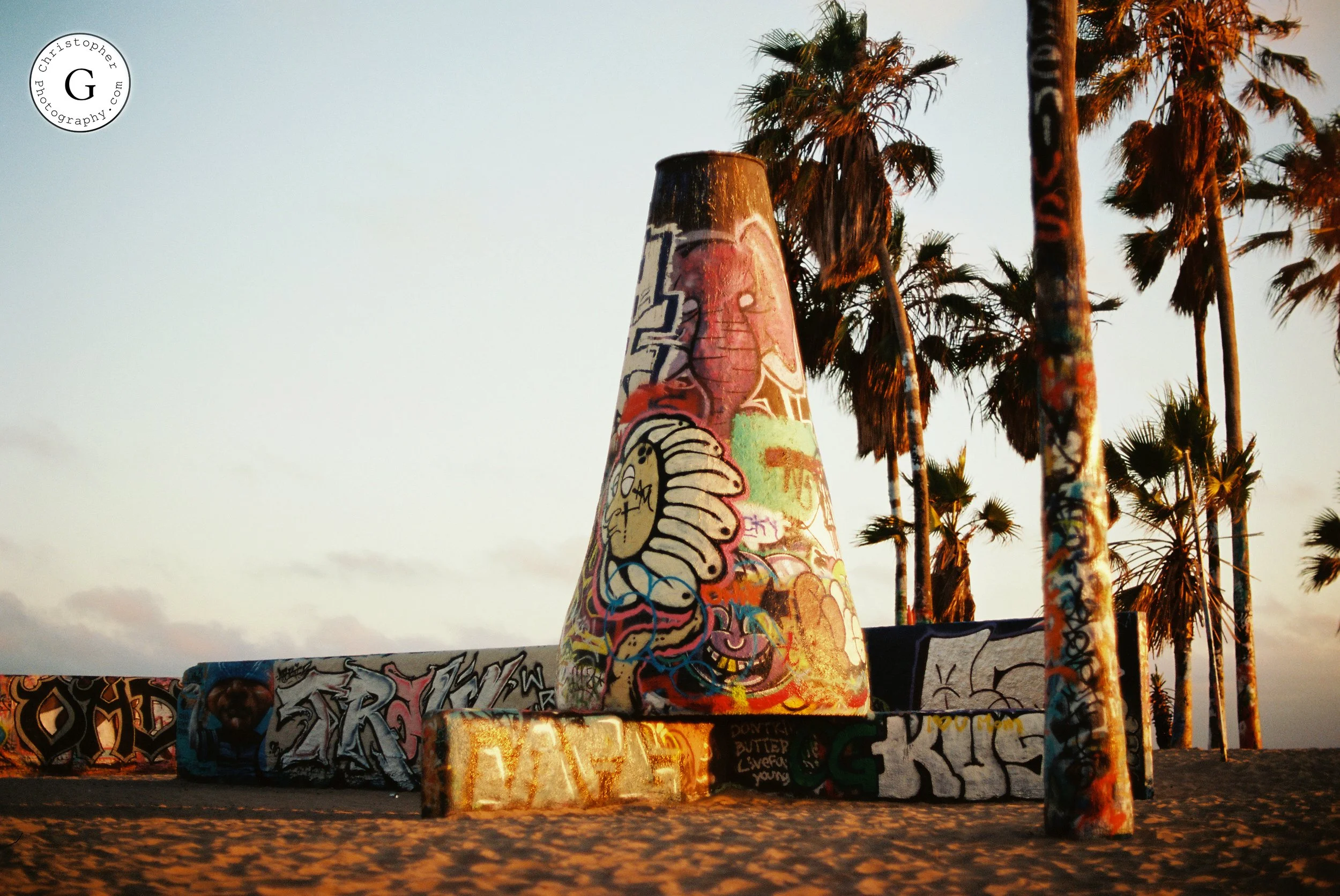 A graffiti-covered cone-shaped sculpture on a platform with palm trees in the background at Venice Beach during sunset shot on 35mm film.