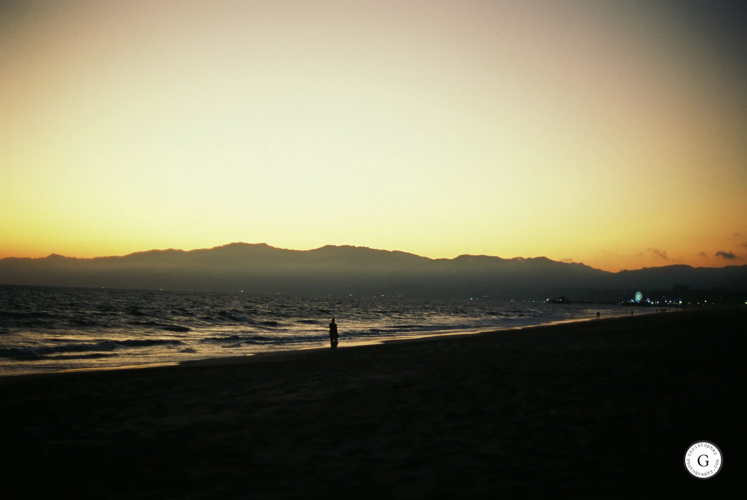A person standing alone on Venice Beach at sunset, with dark sand and the ocean waves, distant mountains on the horizon, and a faint amusement park Ferris wheel lit up in the distance shot on 35mm film.