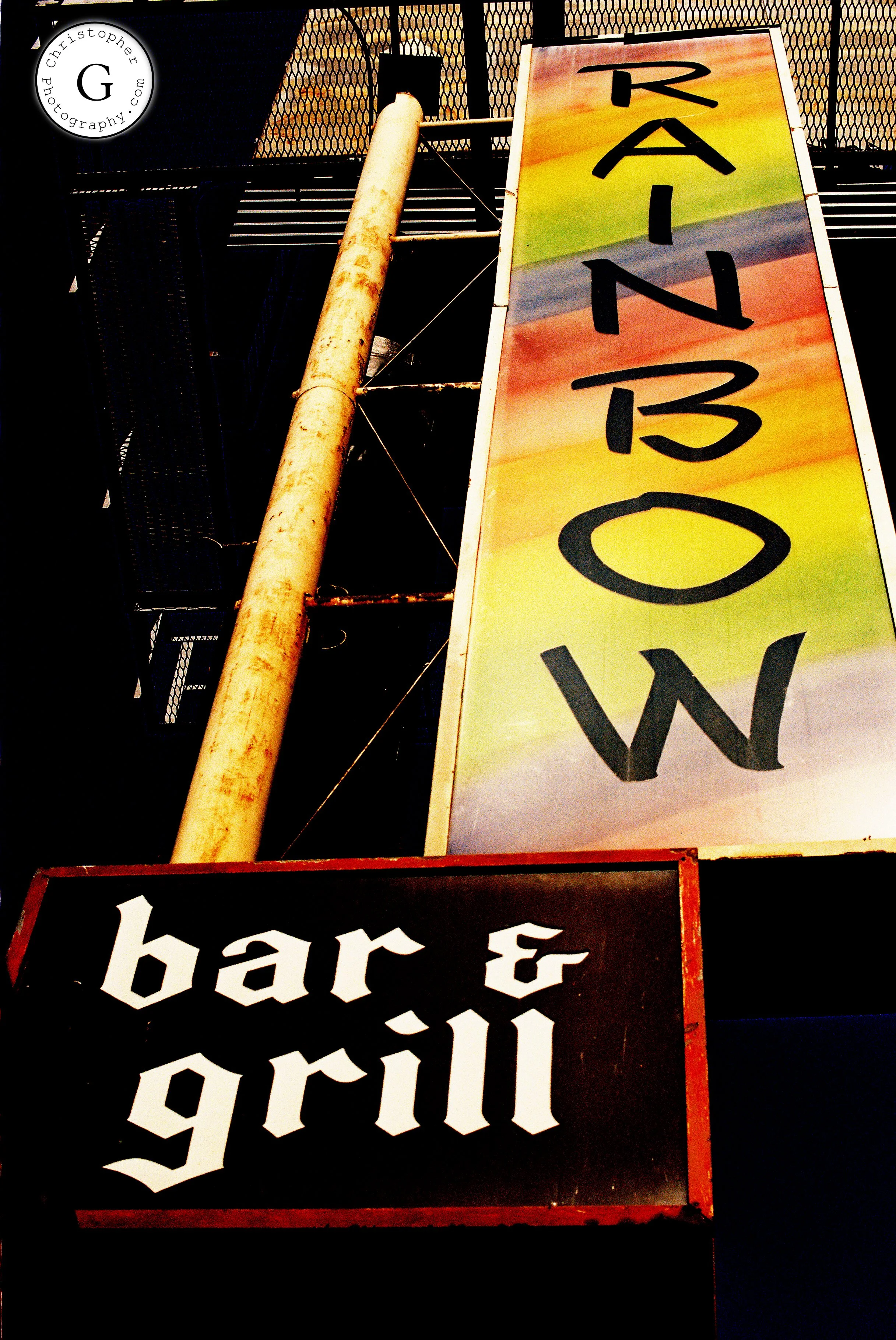 A colorful vertical sign reading 'RAINBOW' and a black sign with white text that says 'bar & grill' underneath shot on 35mm film.