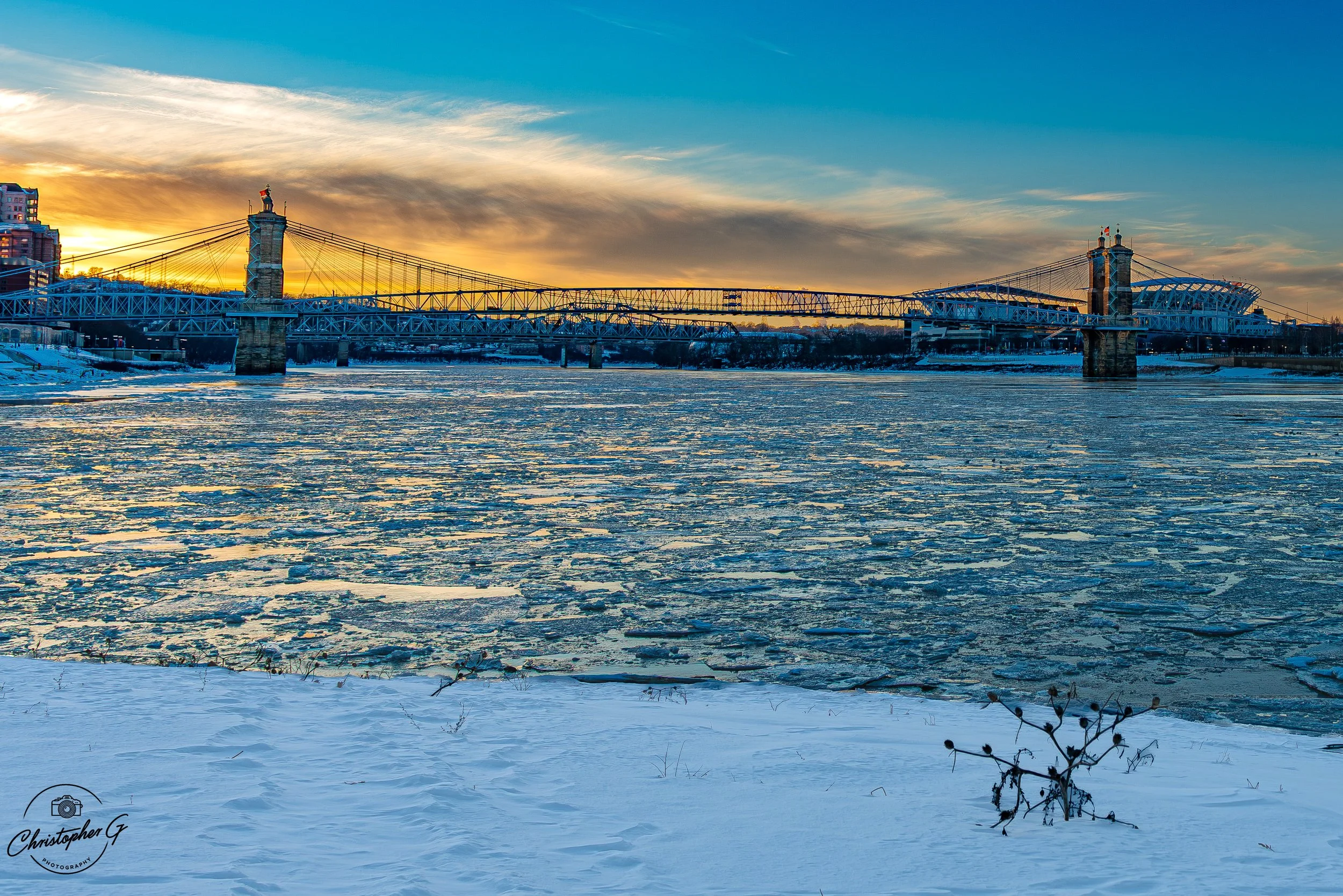 Roebling suspension bridge over a frozen river at sunset with snow-covered ground and partly cloudy sky.