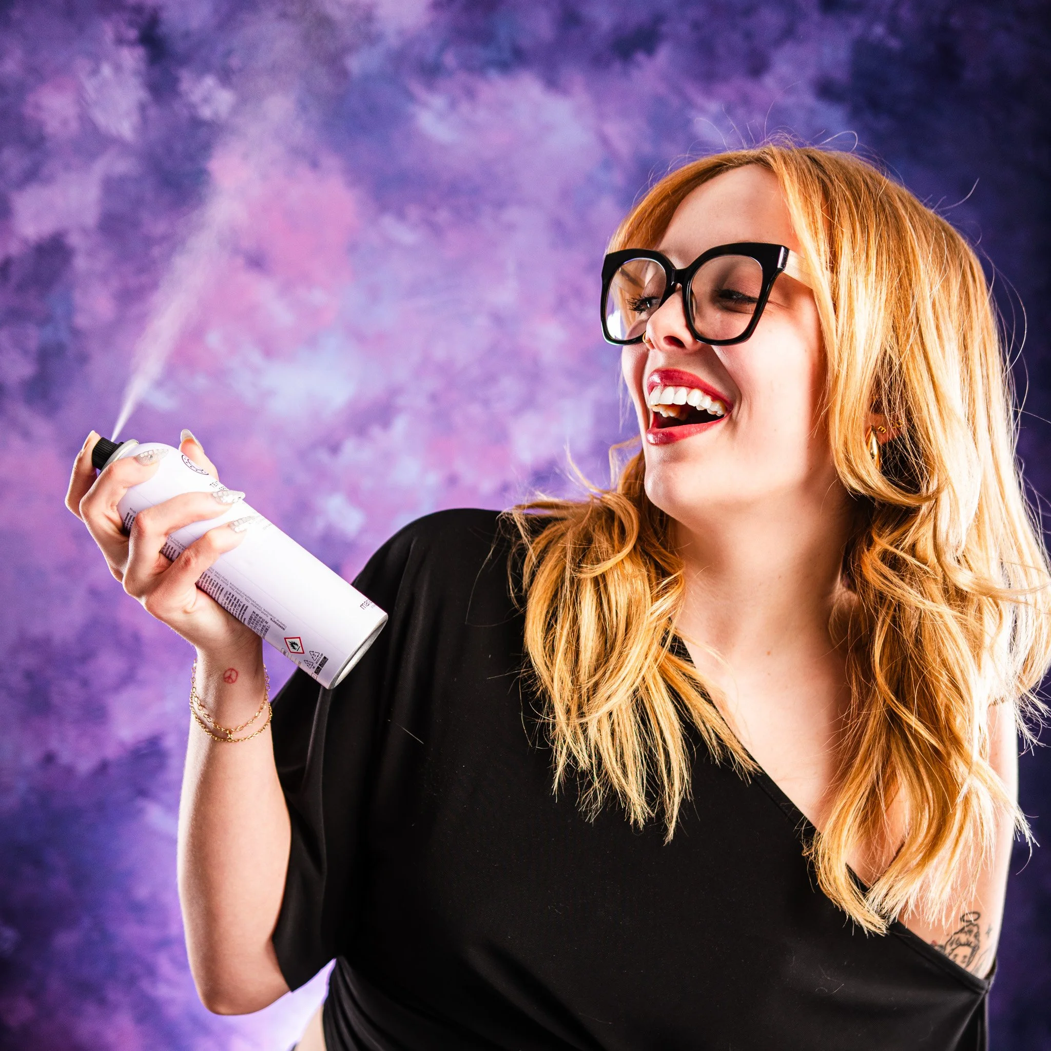 Headshot client for beauty studio having fun with the photo shoot spraying a can and smiling, with colorful purple and blue background.