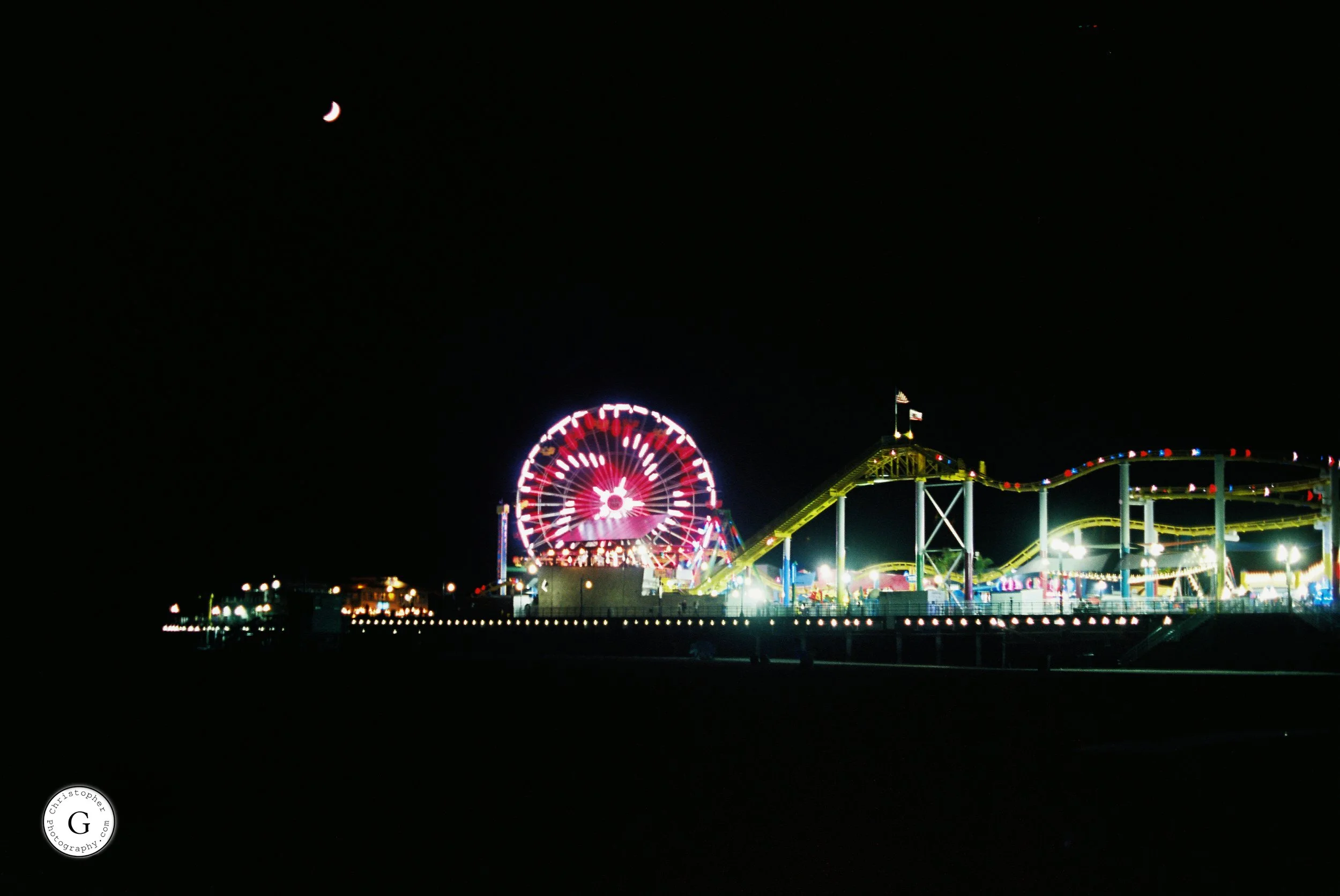 Nighttime view of a brightly lit Pacific Park with a giant Ferris wheel, roller coaster, and other rides against a dark sky with a crescent moon shot on 35mm film .