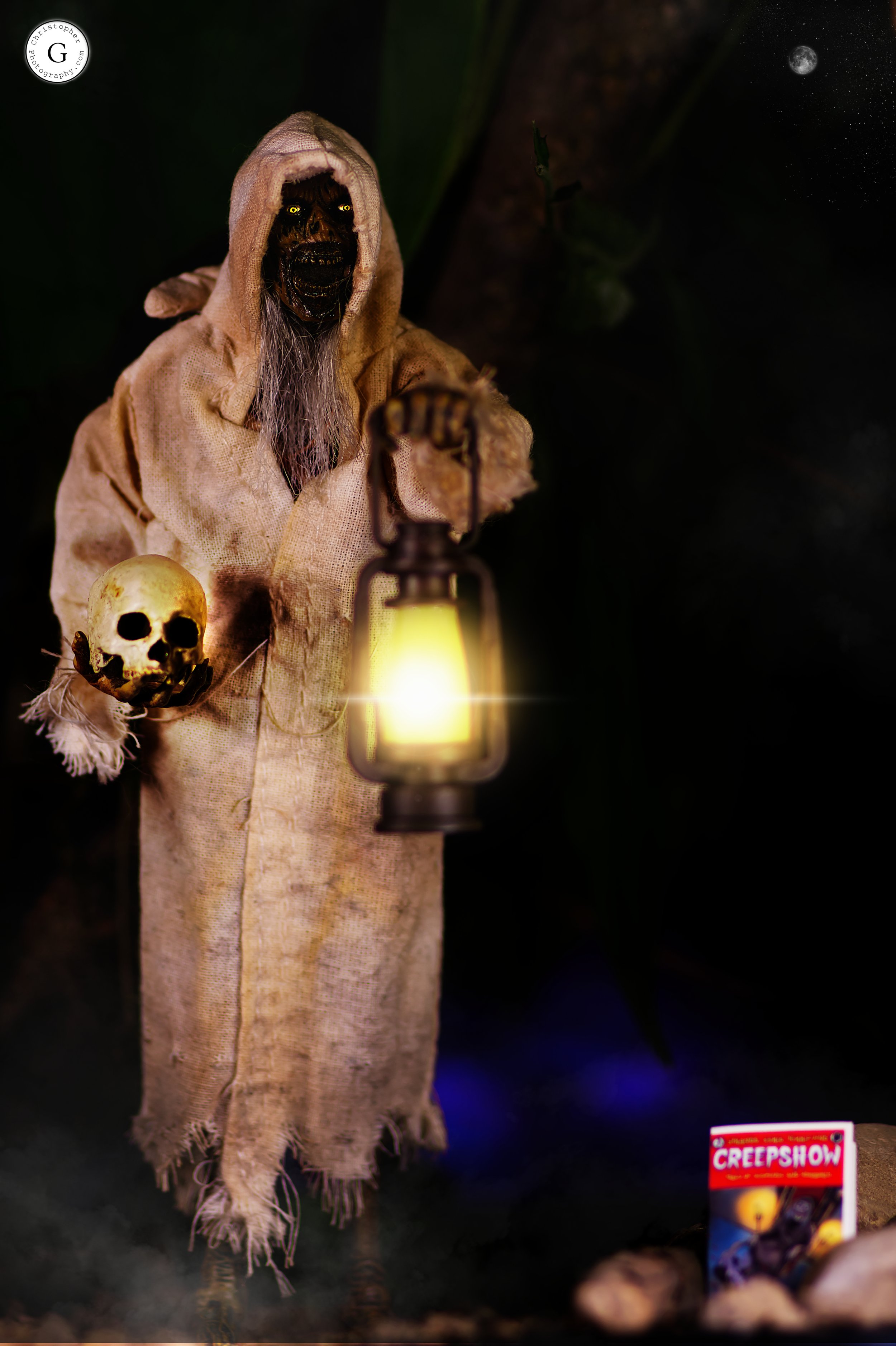 A Halloween decoration of a sinister hooded figure with glowing eyes, holding a skull in one hand and a lantern in the other, standing in a dark setting with a moon in the sky and a magazine titled 'Creepshow' on the ground.