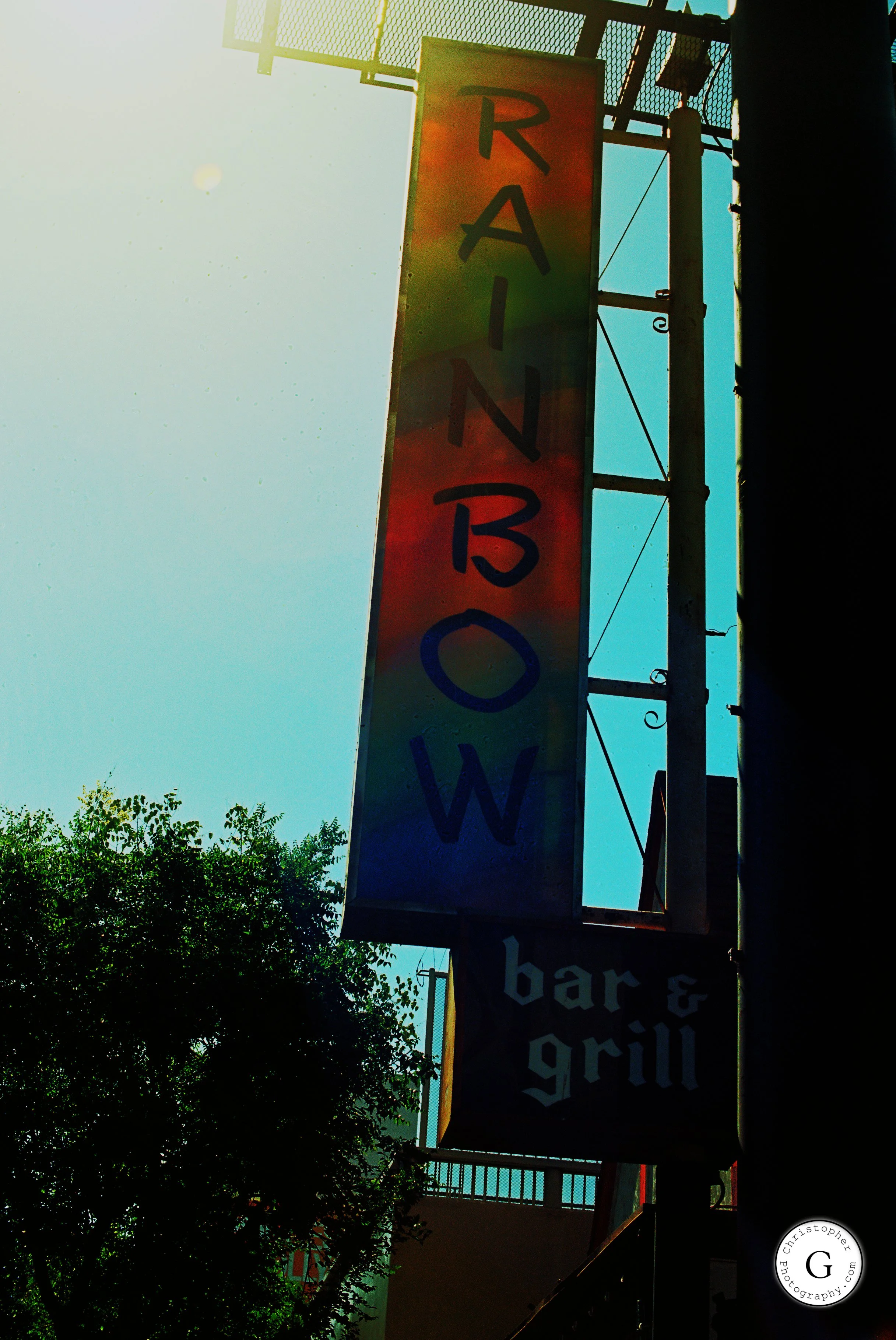 Colorful vertical sign reading 'RAINBOW' with a smaller sign below that says 'bar & grill', against a bright sky with trees at the bottom left shot on 35mm film.