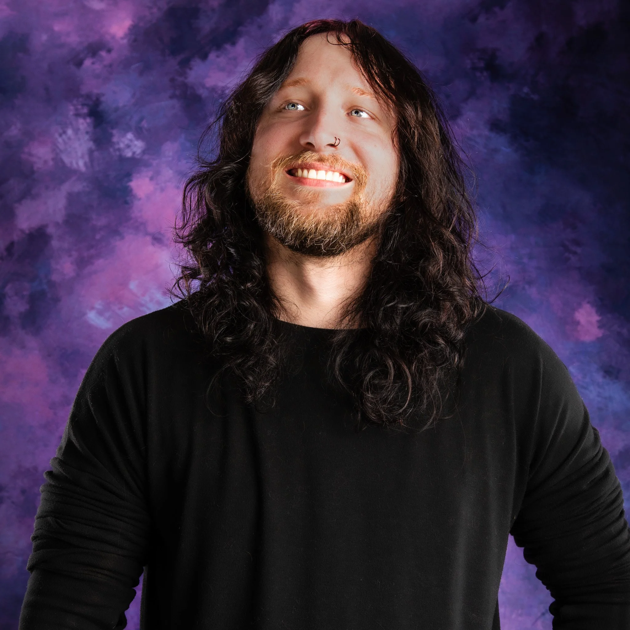 Portrait of a man with long curly hair and a beard, smiling and wearing a black shirt, against a purple and blue patterned background.