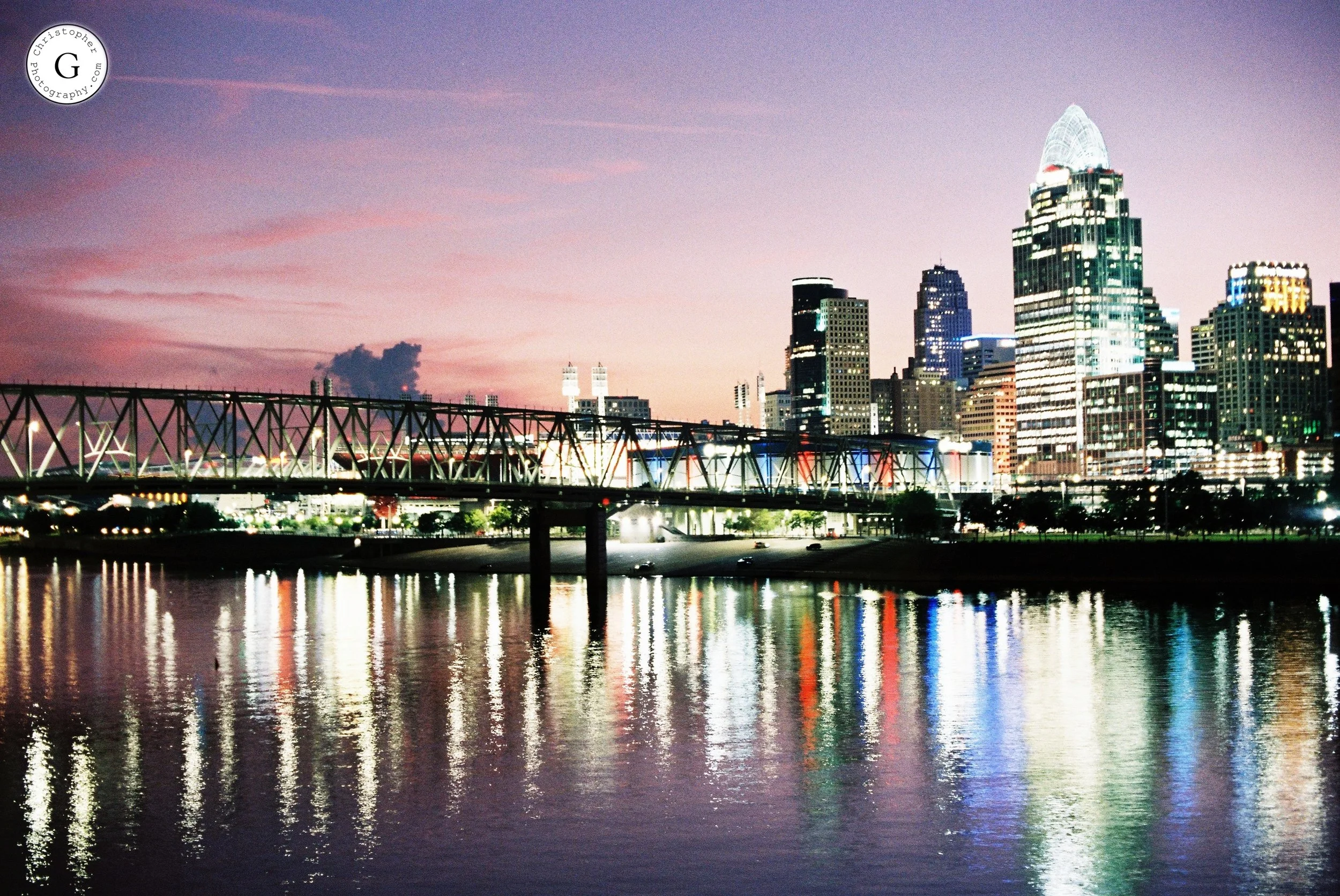 Cincinnati skyline at dusk with lit buildings and reflections on the river, including a bridge in the foreground shot on 35mm film 