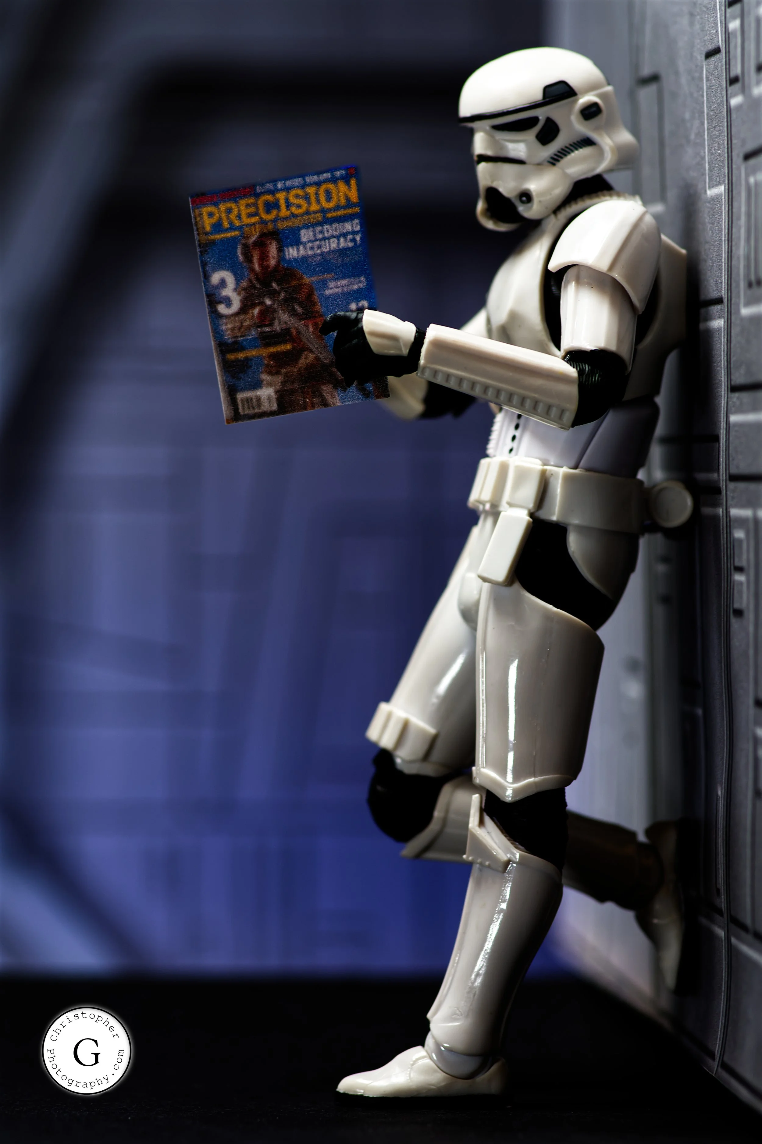 A plastic Stormtrooper toy leaning against a wall while reading a magazine titled 'Precision' with a woman on the cover. The background is blurred, and the toy appears to be in a toy or display setting.