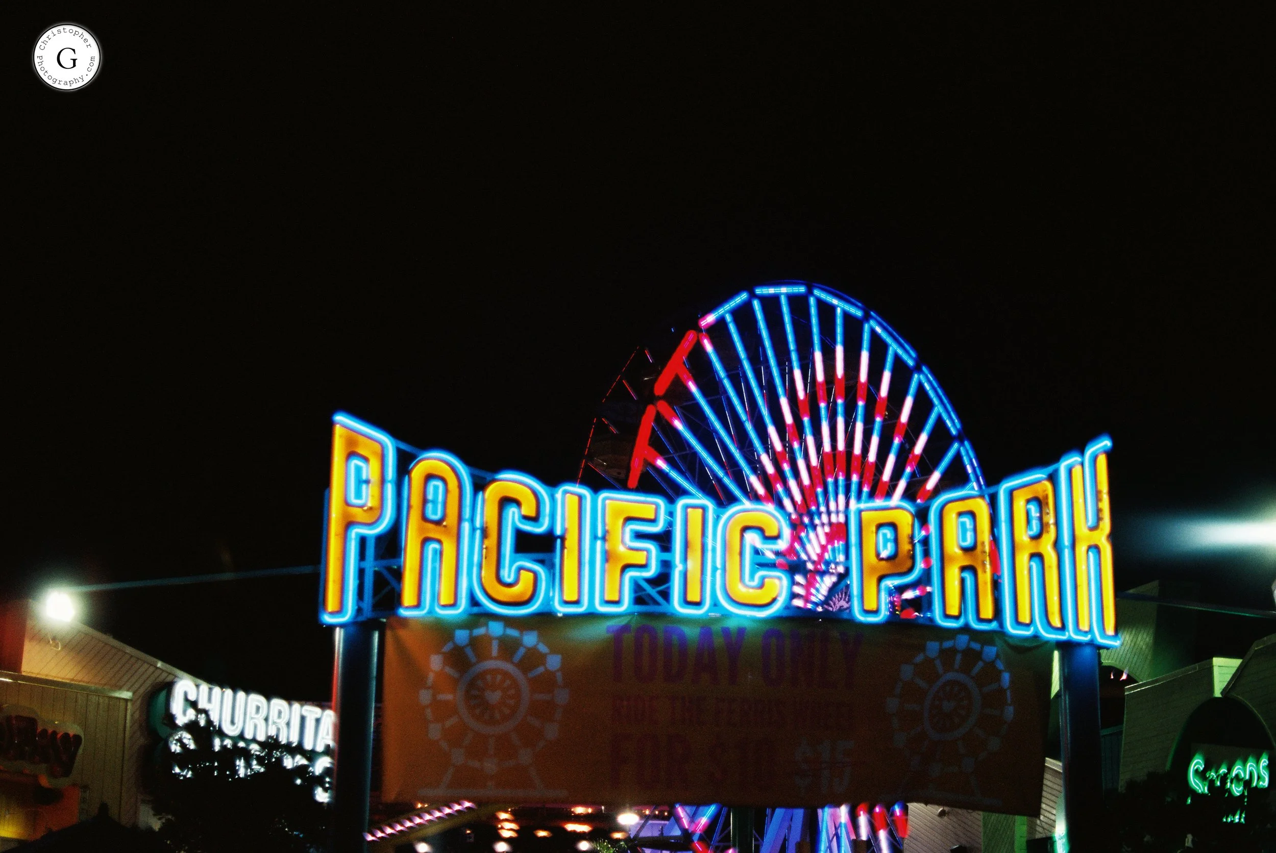 Night view of a neon-lit sign for Pacific Park amusement park with a Ferris wheel in the background shot on 35mm film.