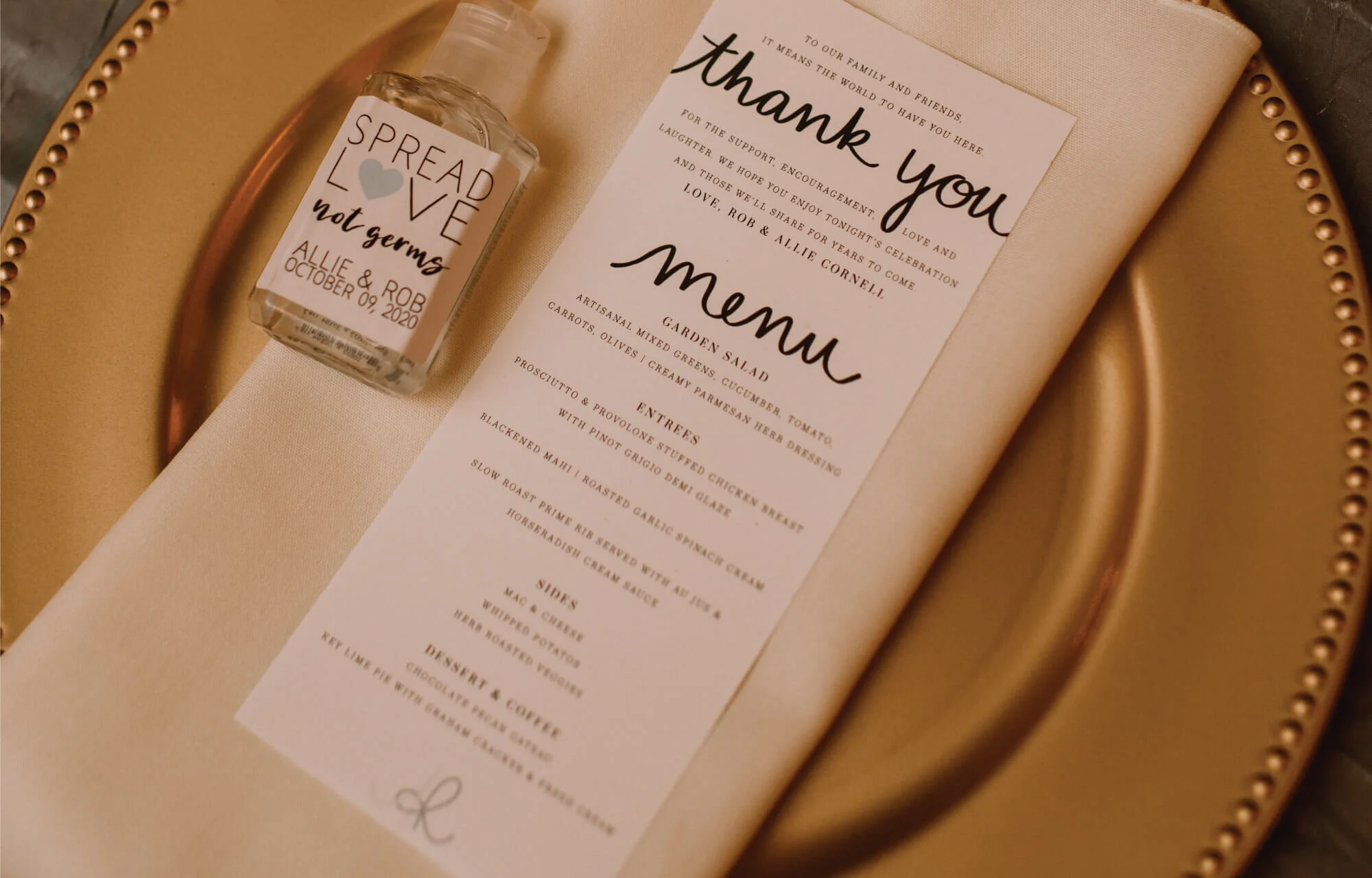 Thank you card and menu laying on a cream colored dinner napkin on a gold charger with hand sanitizer next to it that says spread love not germs