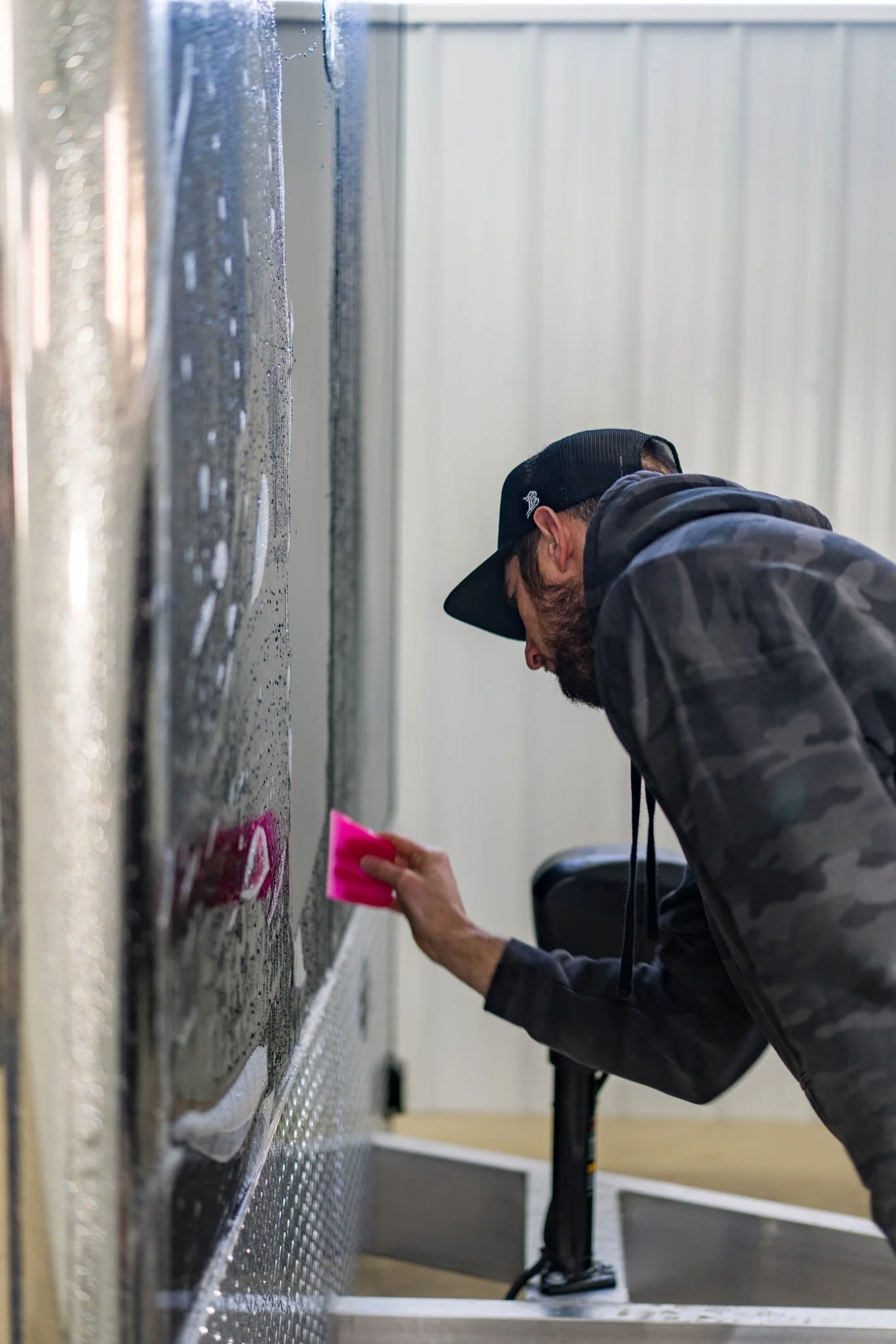 A man wearing a black cap and camouflage hoodie is cleaning or inspecting a large metallic surface, possibly an industrial or workshop setting. He is holding a pink cloth or sponge, and the surface has visible water droplets or condensation.