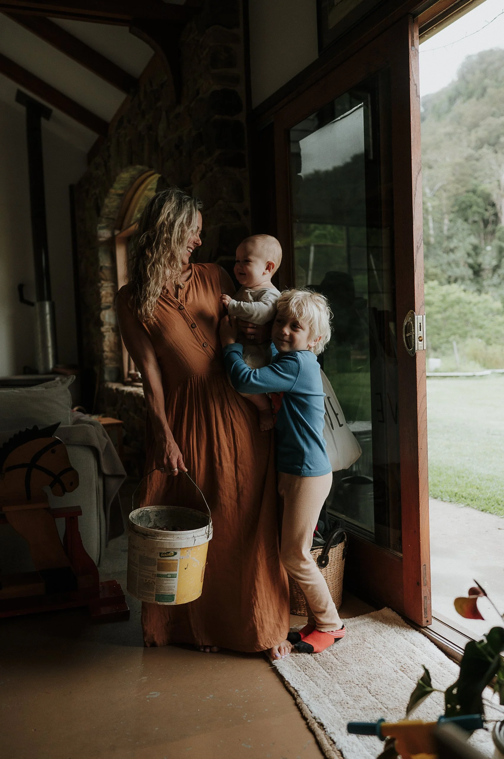 A woman with curly blonde hair in a rust-colored dress playing with two young children near a large open door inside a house.